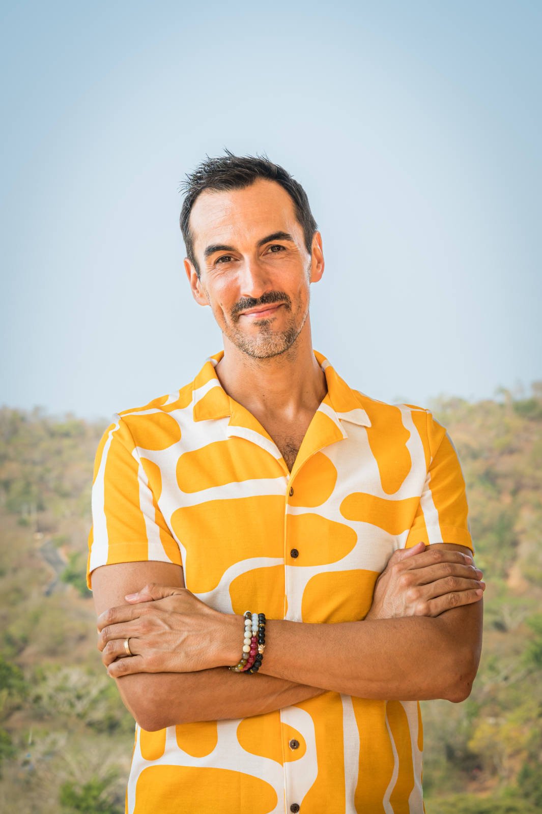 Man in yellow patterned shirt standing with arms crossed outside, blue sky background.