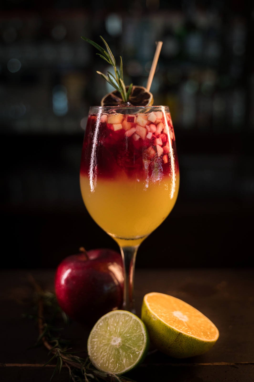 A fruity cocktail in a wine glass, garnished with diced fruit, rosemary, and a lime wheel. Surrounding the drink are a whole apple, a half lime, and a half orange.