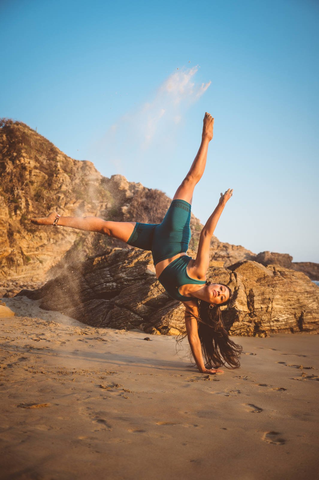 Person performing a handstand on a sandy beach with rocky cliffs in the background, wearing teal athletic wear.