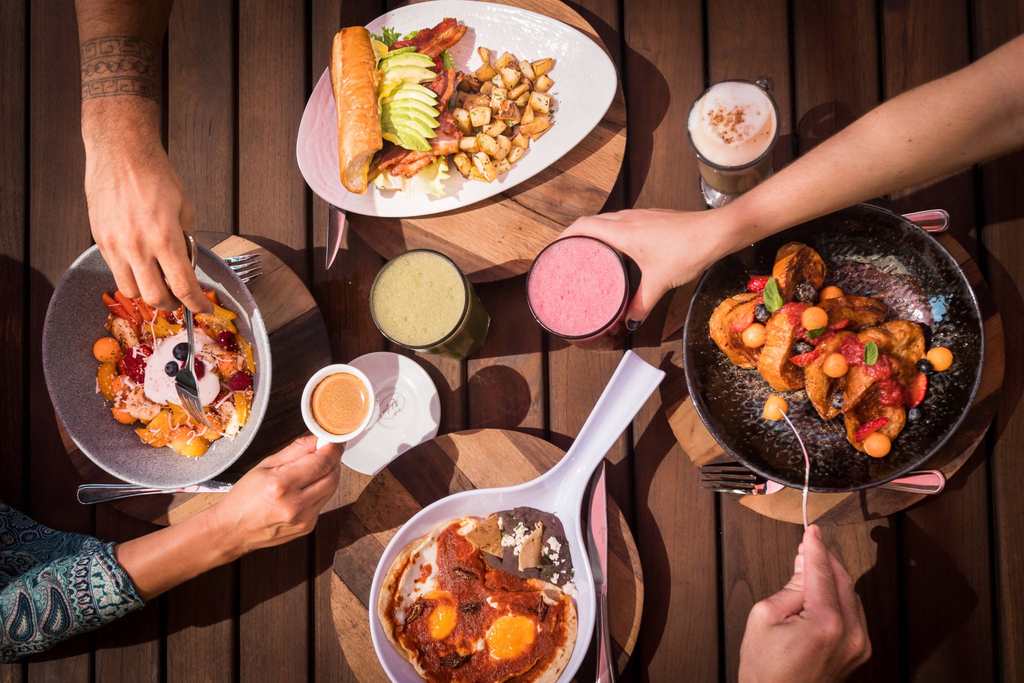 Overhead view of a brunch table setting with various dishes including toast with avocado and bacon, fruit bowls, sunny-side-up eggs, roasted potatoes, and beverages like smoothies and espresso.