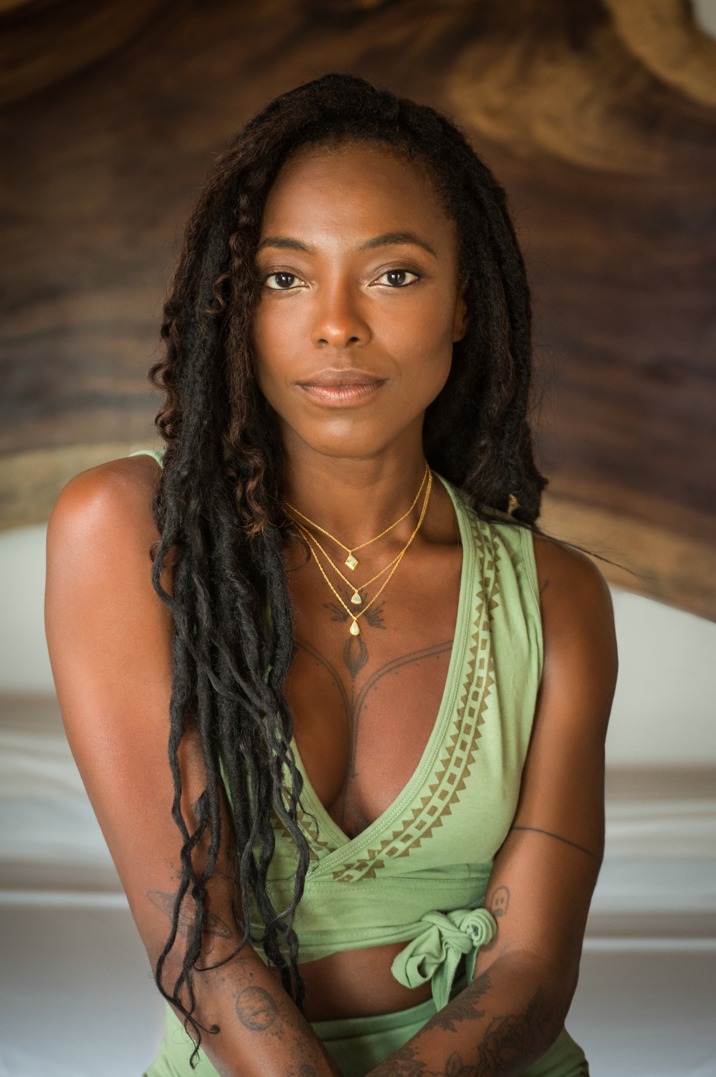 Person with long dreadlocks, wearing green clothing and layered necklaces, sitting with a neutral expression.