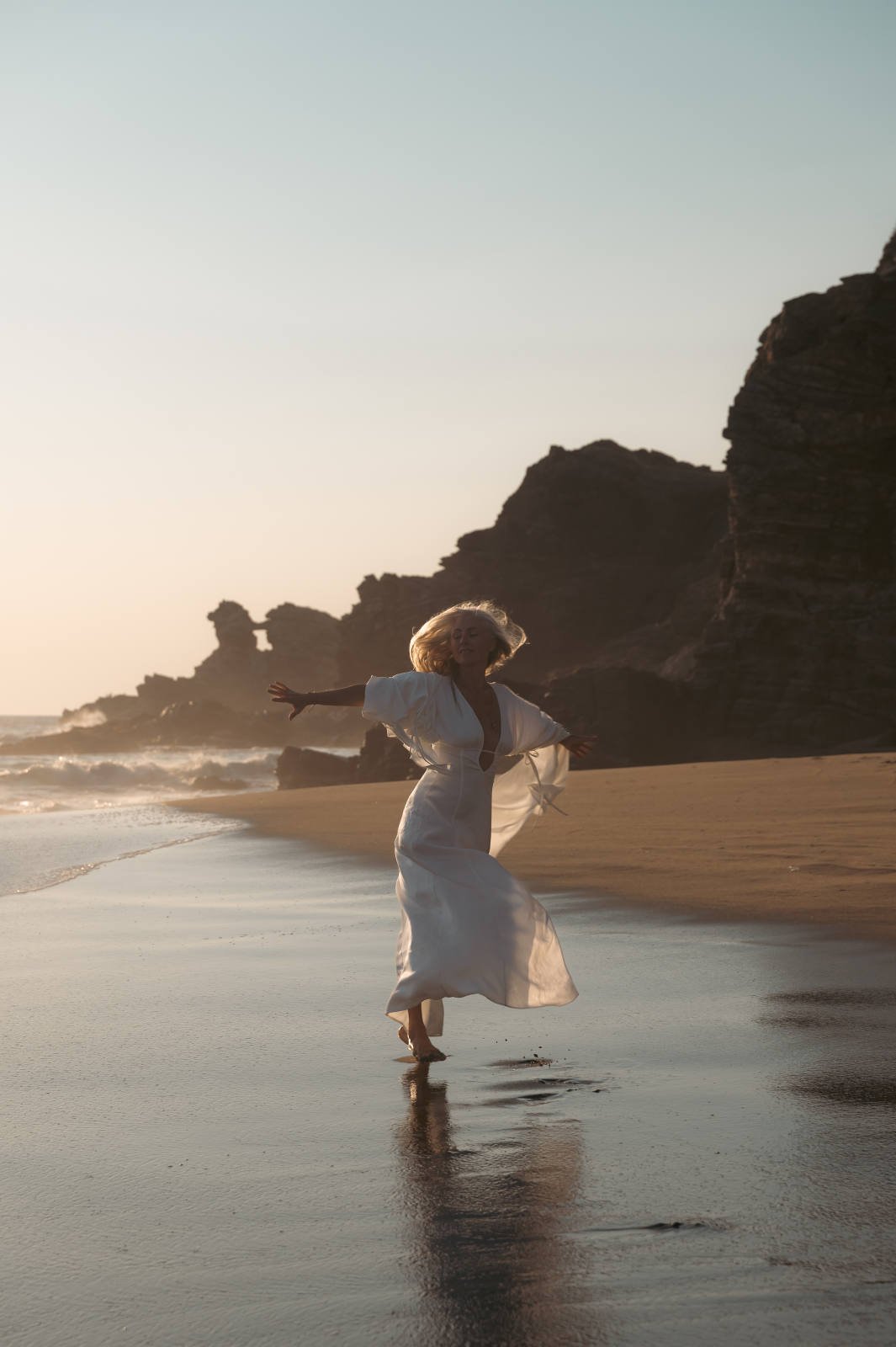 Woman in white dress dancing on a beach at sunset with rocky cliffs in the background.