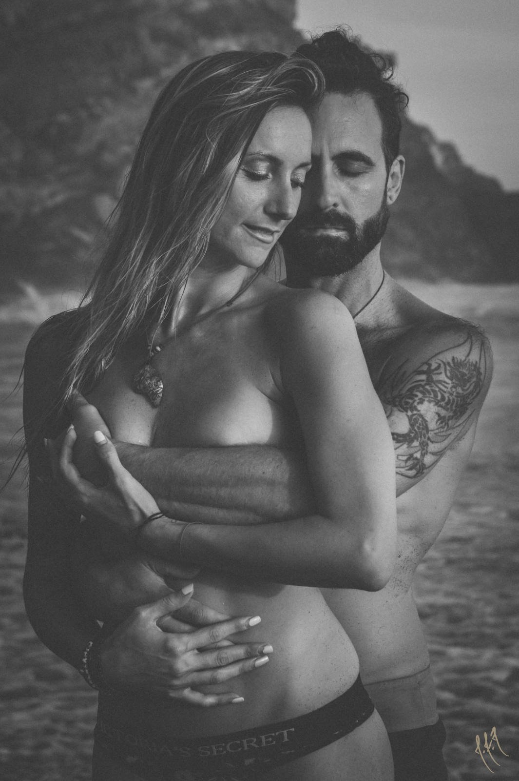 Black and white photo of a couple embracing on a beach, the man has a tattoo on his arm.