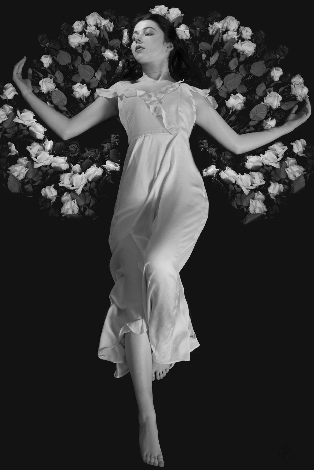 Black and white photo of a woman in a flowing white dress, lying on a bed of roses with her arms outstretched.