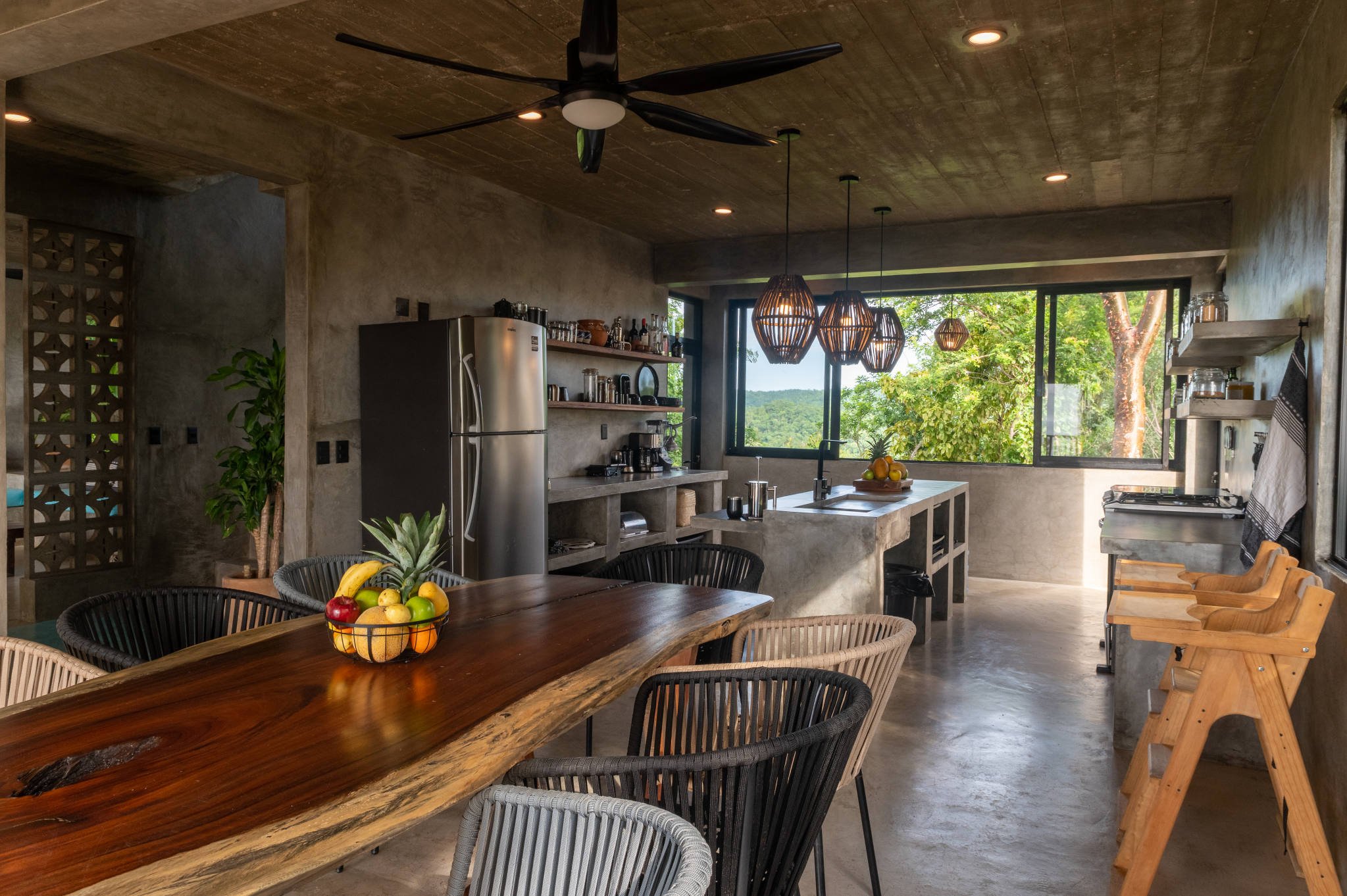 Modern kitchen with wooden table and chairs, stainless steel appliances, concrete countertops, pendant lights, ceiling fan, fruit bowl, open shelving, and large window with view of trees.