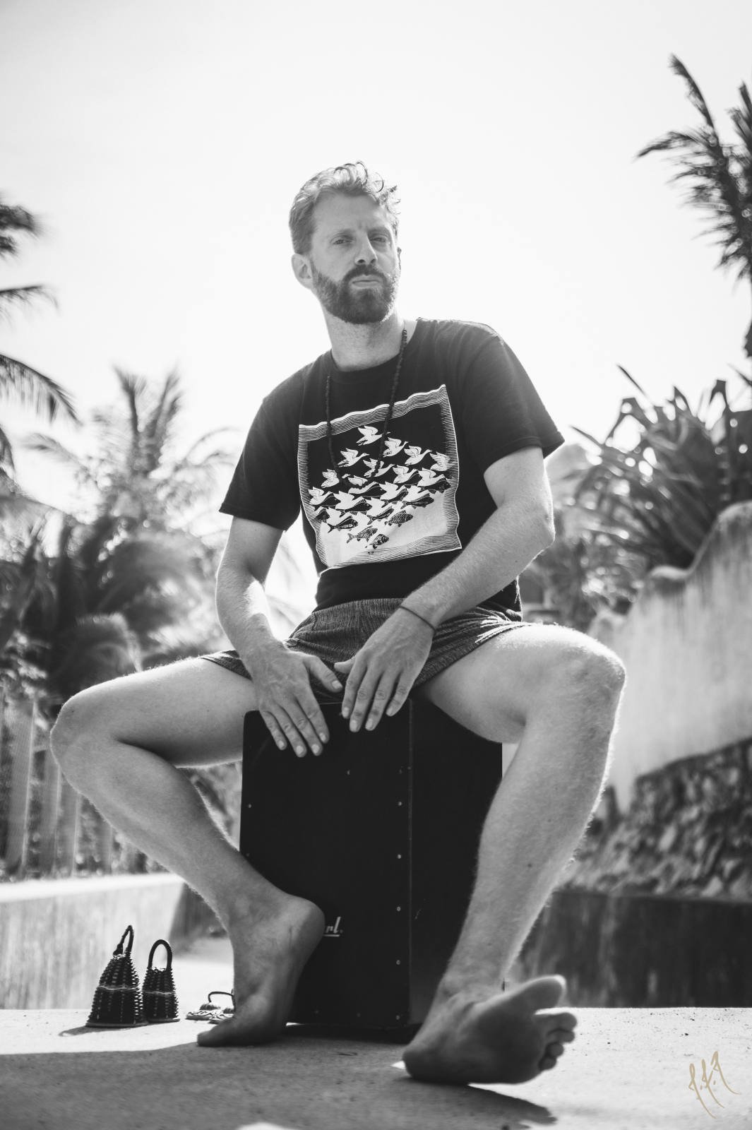 A man sitting outdoors on a cajón, playing the instrument. He is barefoot and wearing a black t-shirt with a bird pattern and shorts. Tropical plants and trees are in the background.