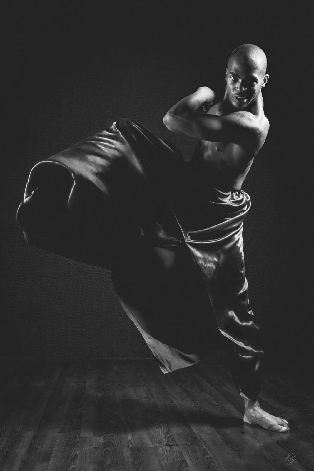 Black and white photo of a dancer performing a dramatic pose with a flowing fabric, bare-chested, on a wooden floor.