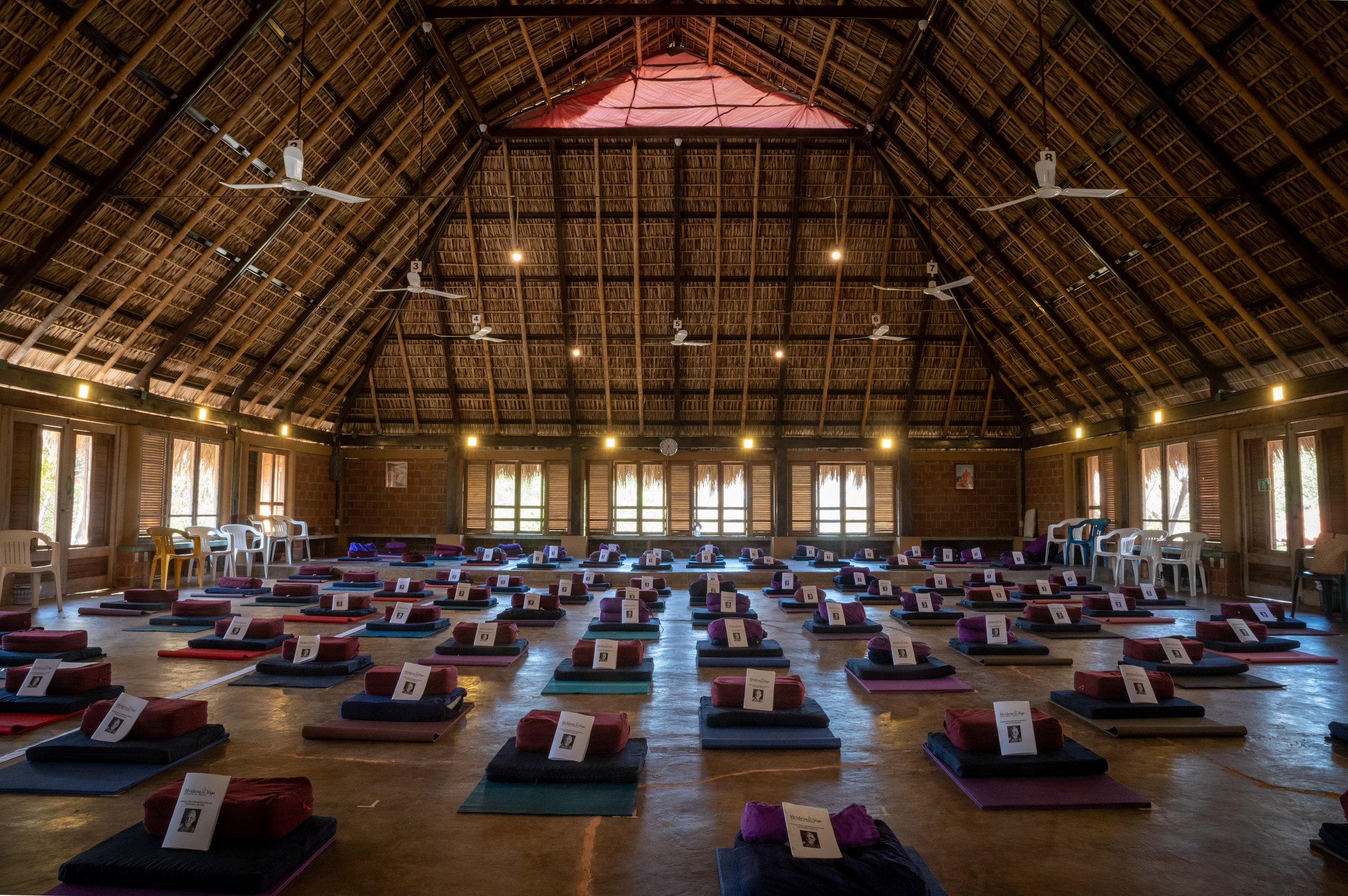 Interior of a meditation hall with arranged mats and cushions on the floor, under a high thatched ceiling.