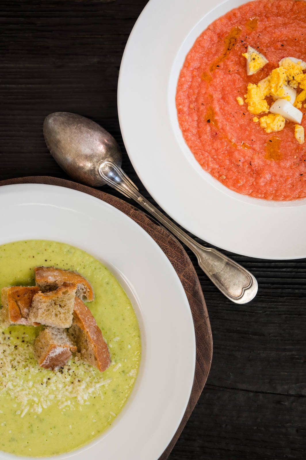 Plates of red and green creamy soups with croutons and egg garnishes, served with a silver spoon on a dark wooden surface.