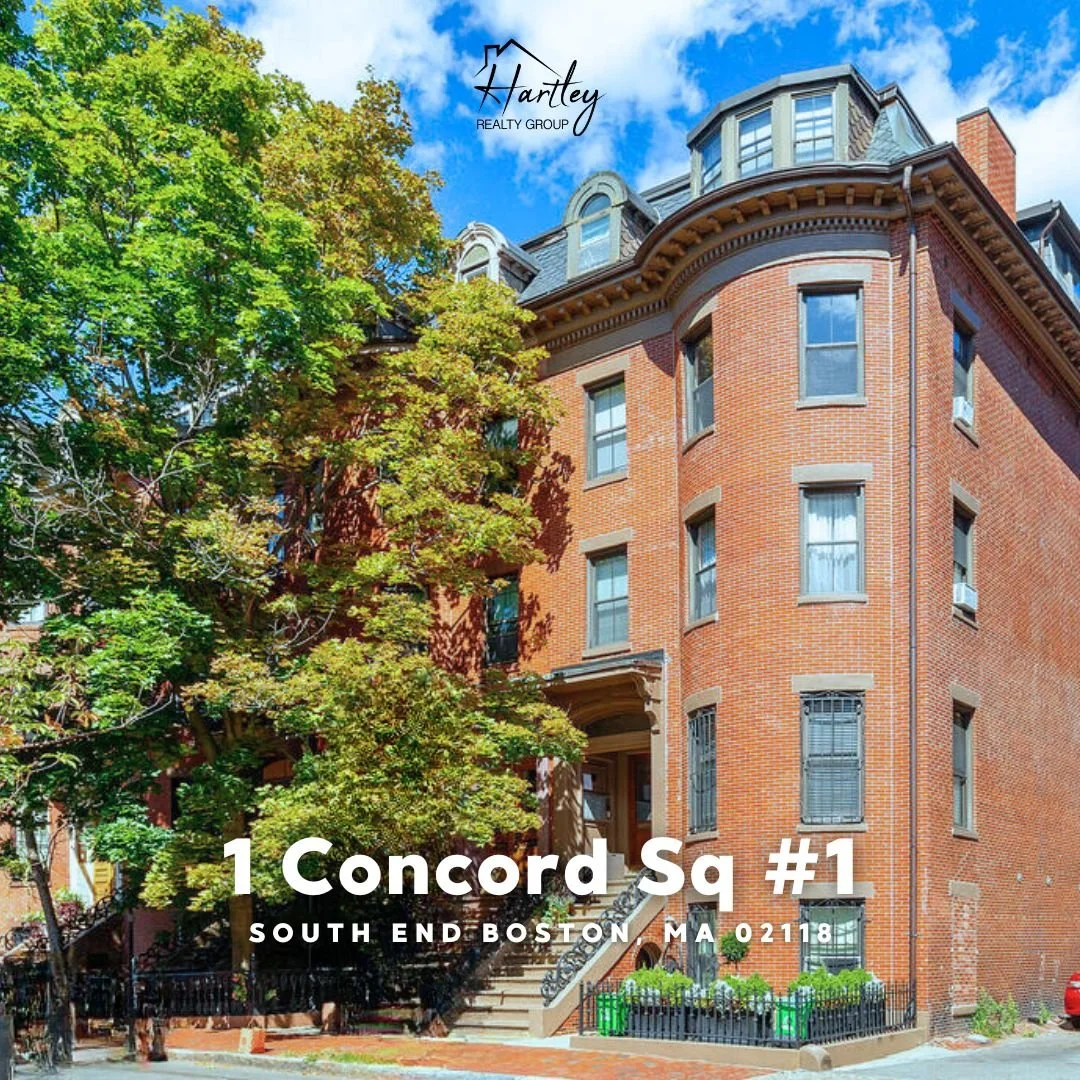 A red brick corner building with multiple windows and a rounded rooftop in South End Boston, Massachusetts, under a partly cloudy sky, with a large tree partially covering the building, a black fence with planters, and steps leading to the entrance.