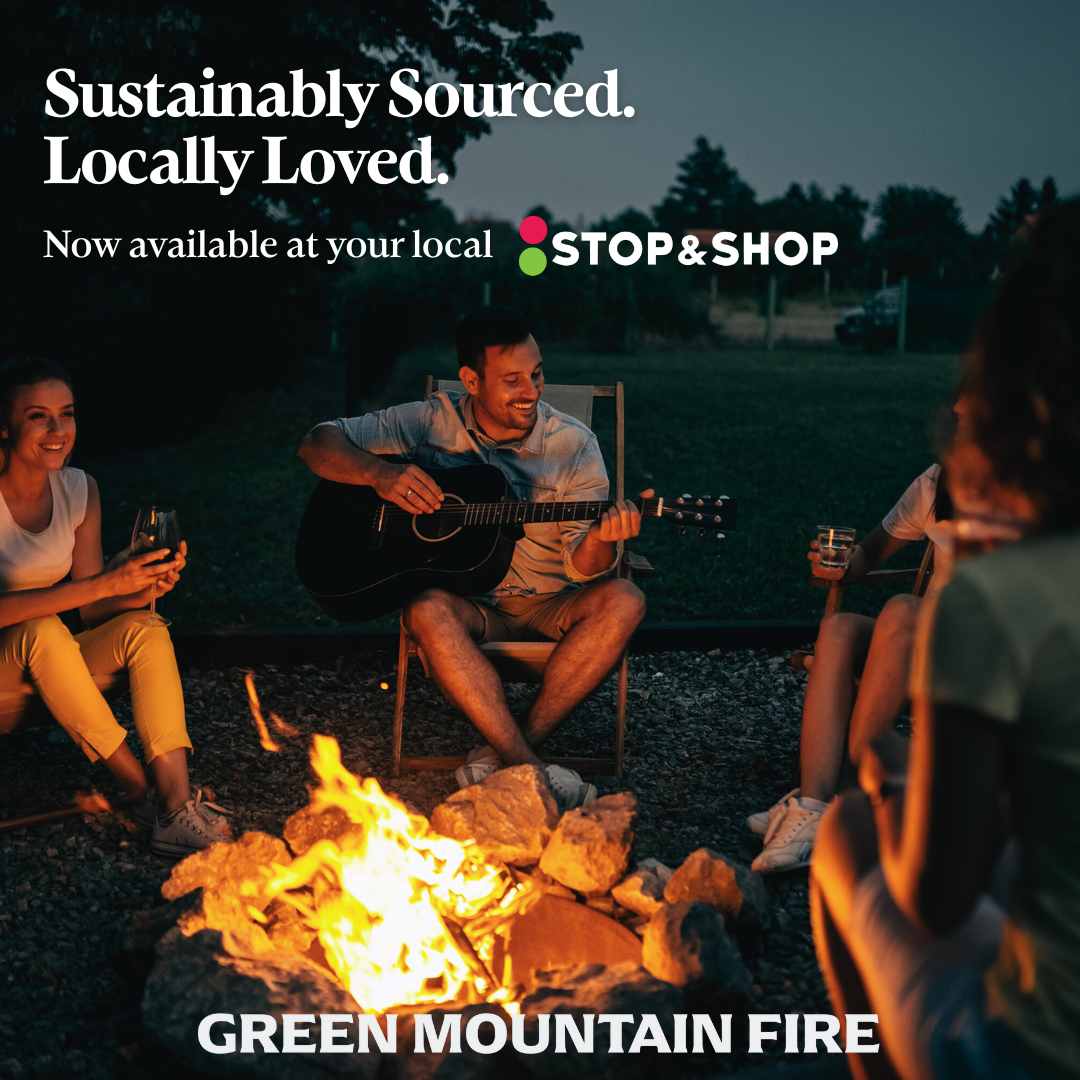 Group of friends sitting around a campfire at night, one playing guitar, others with drinks, outdoors in a green, mountainous area.