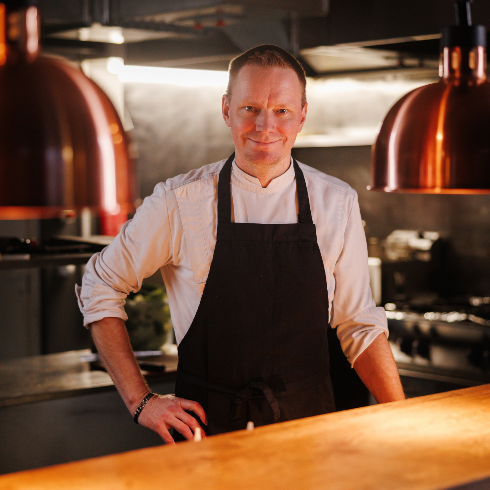A male chef standing in a modern kitchen, wearing a white chef's coat and black apron, smiling, with copper pendant lights hanging above a wooden counter.