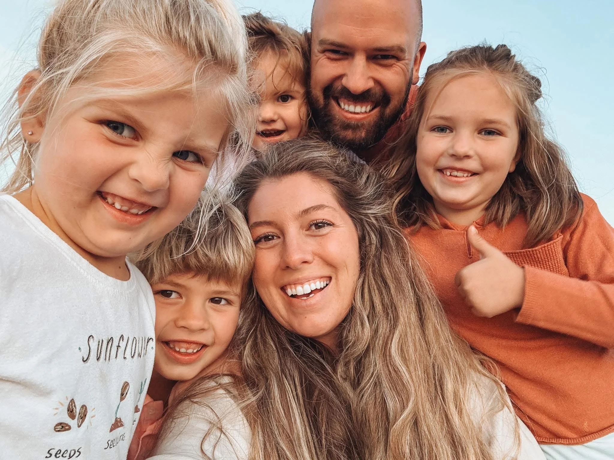 A family of six smiling and taking a selfie outdoors, with a clear blue sky background.