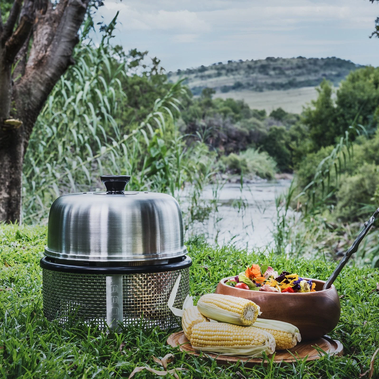 Outdoor picnic setup with a metal covered food warmer, a ceramic bowl with fresh salad, and ears of corn on a wooden tray by a river with lush trees and mountains in the background.