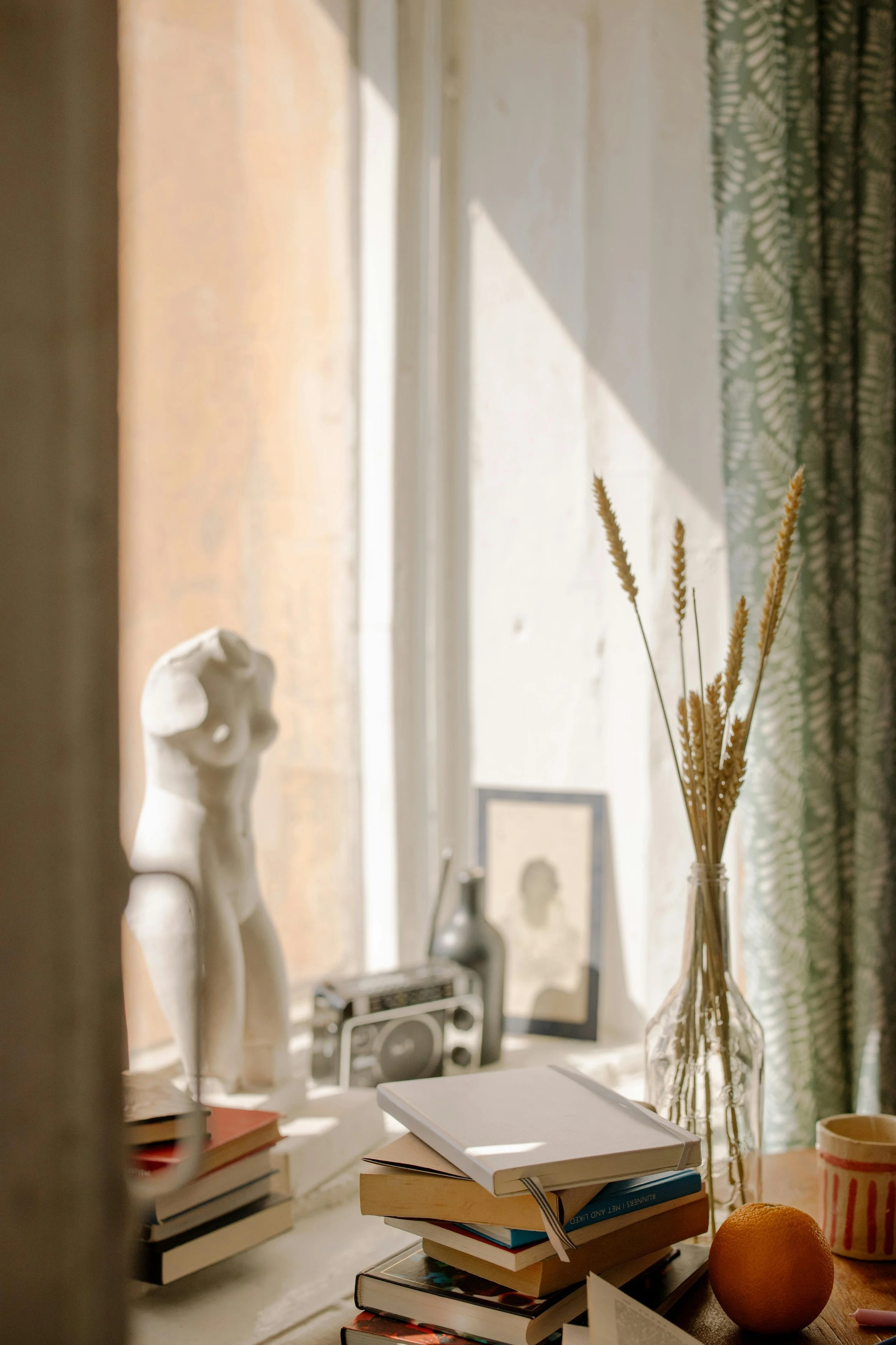 A windowsill with a statue, books, a framed photo, a vintage radio, and a vase with dried grasses, near a window with patterned curtains and sunlight streaming in.