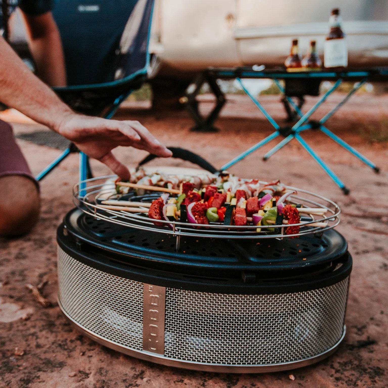 Person grilling skewers of vegetables and meat on a portable outdoor grill.
