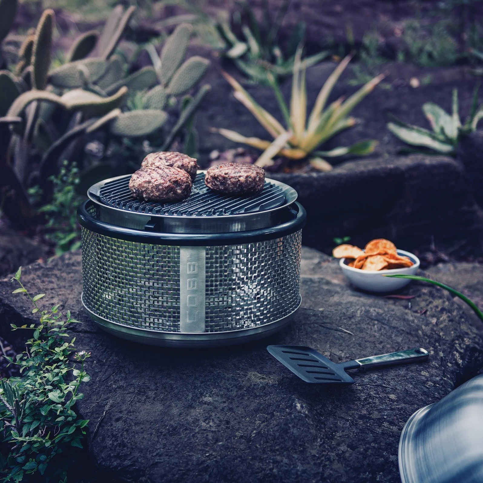 Portable grill with three burger patties cooking on a stone surface outdoors, surrounded by drought-tolerant plants, with a small bowl of chips and a spatula nearby.