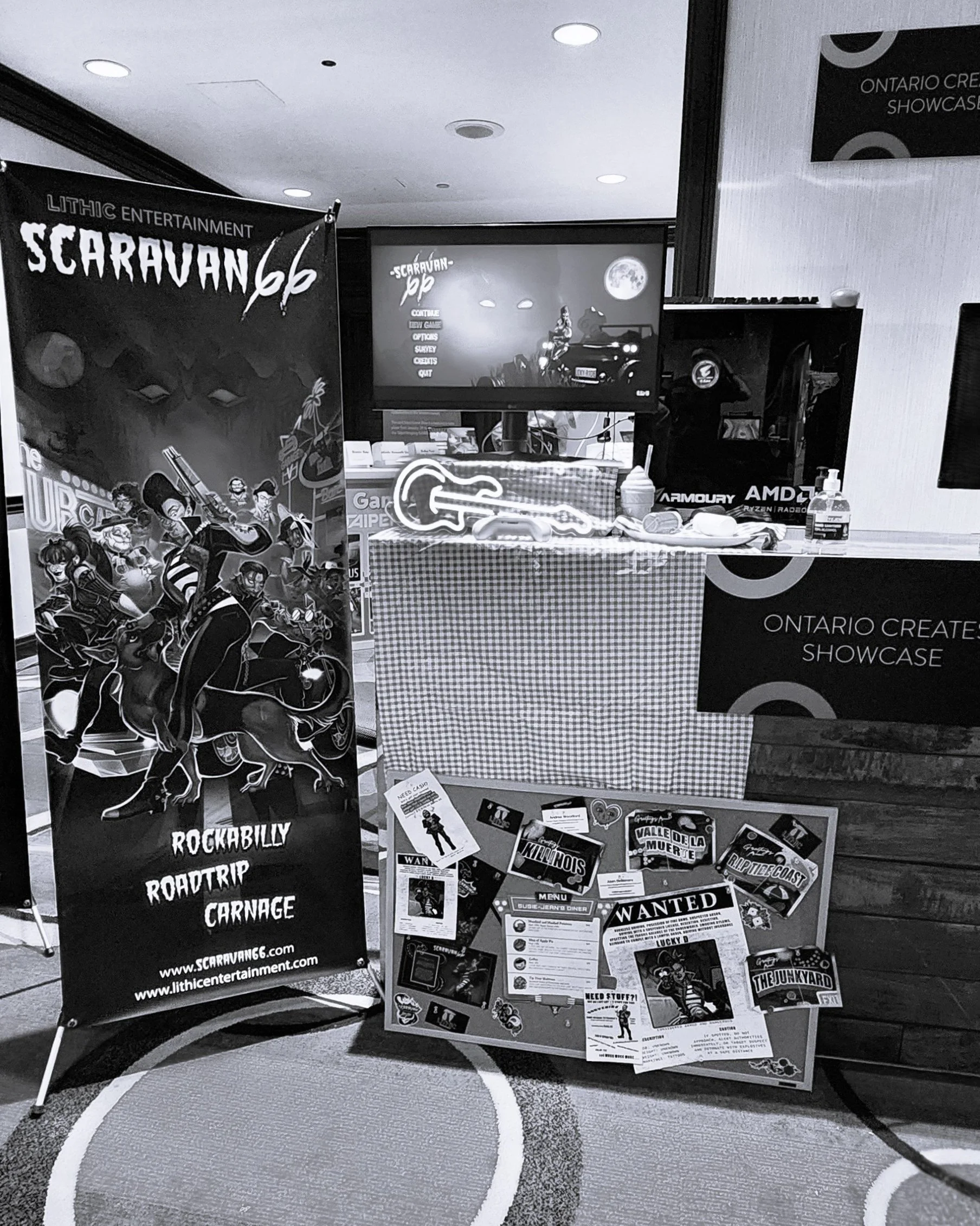 Exhibit setup with promotional banners and boards for Lithic Entertainment's 'Scaravan 66,' featuring a neon guitar sign, a display screen, and various promotional materials.