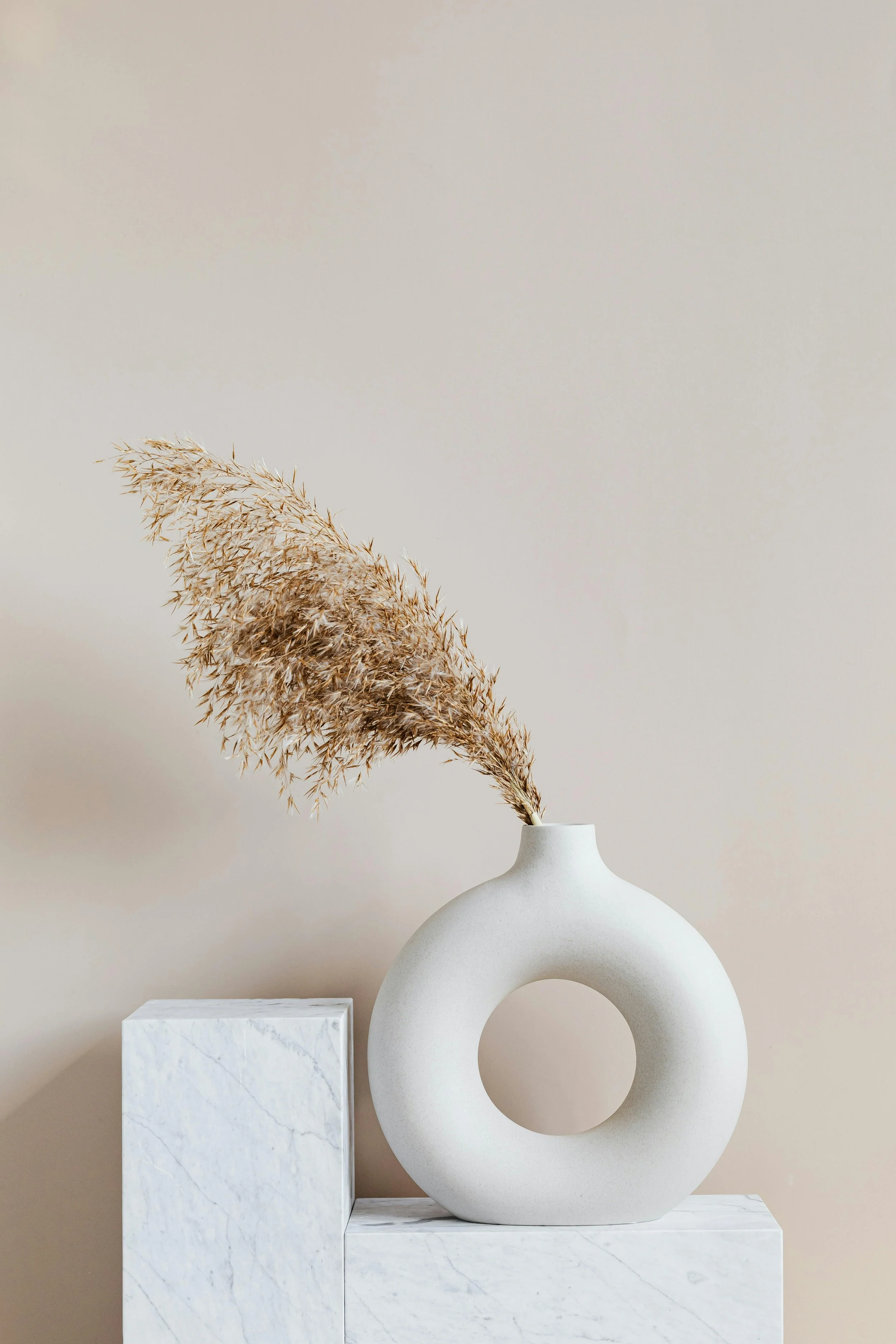 A modern white ceramic vase with a circular hole in the center, holding a large dried pampas grass stem, placed on a white marble pedestal against a neutral beige wall.