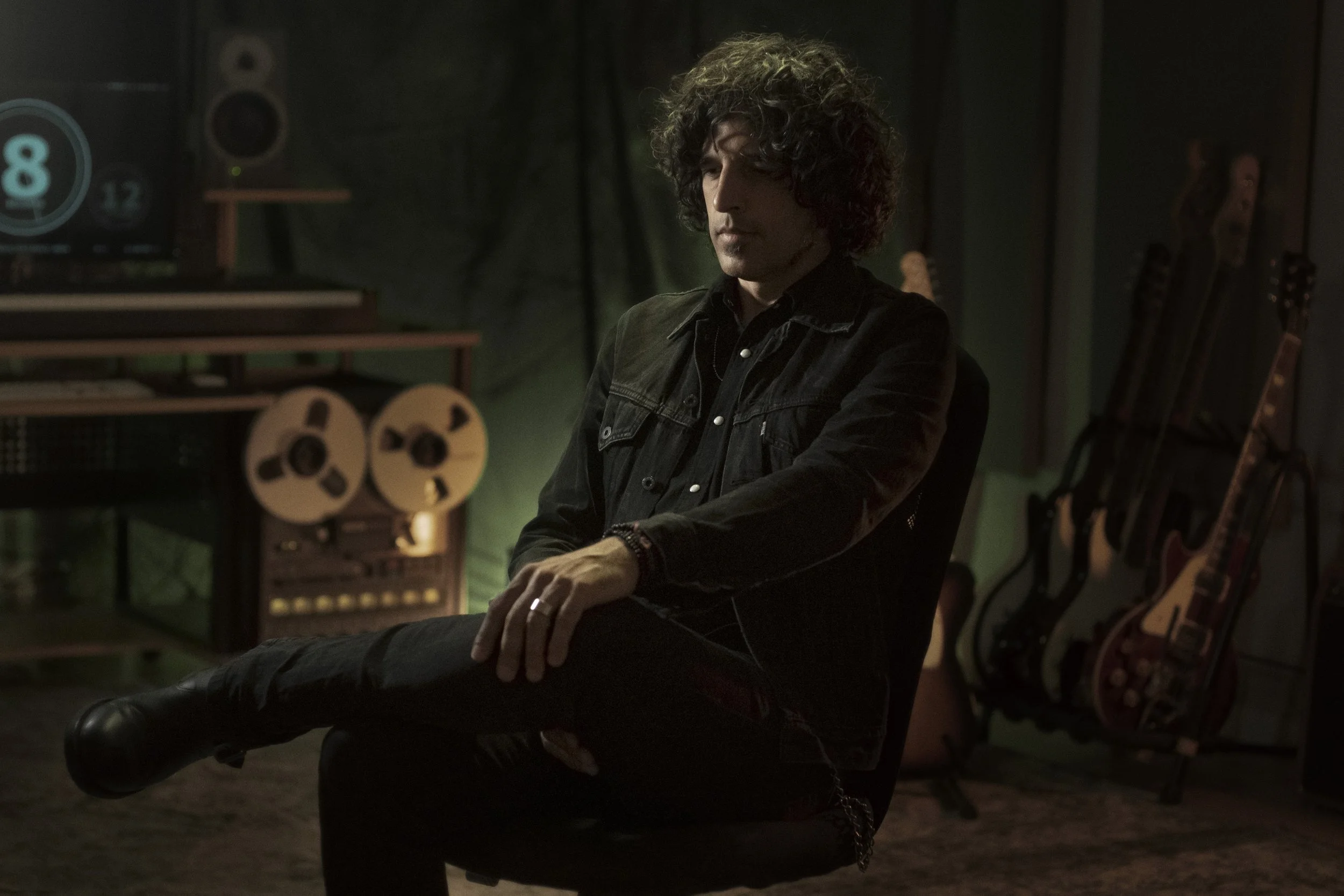Juan Navazo, sentado en una silla en un estudio de grabación con guitarras en el fondo, y una pantalla con un reloj digital a la izquierda.