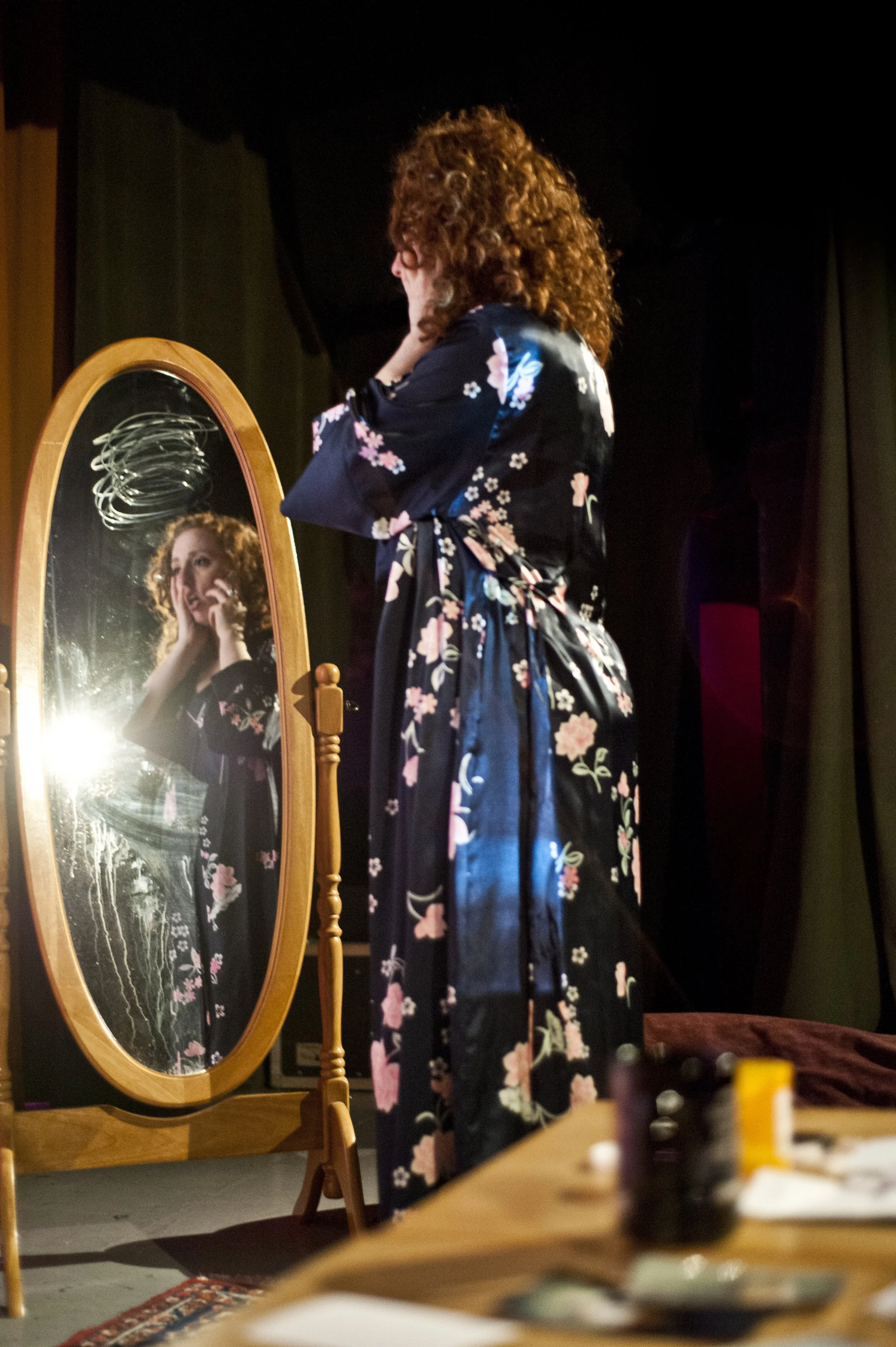 A woman in a floral kimono stands in front of an oval mirror, adjusting her hair. Her reflection is visible in the mirror. A table with various objects is in the foreground.