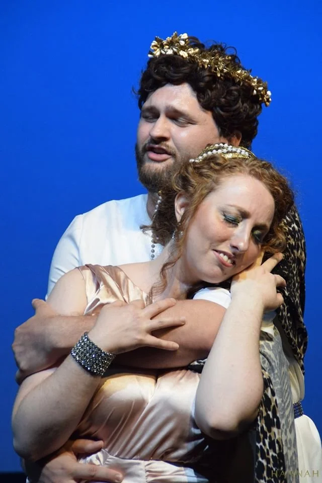 A man and woman dressed in classical attire with crowns stand embracing each other, eyes closed, against a blue background.