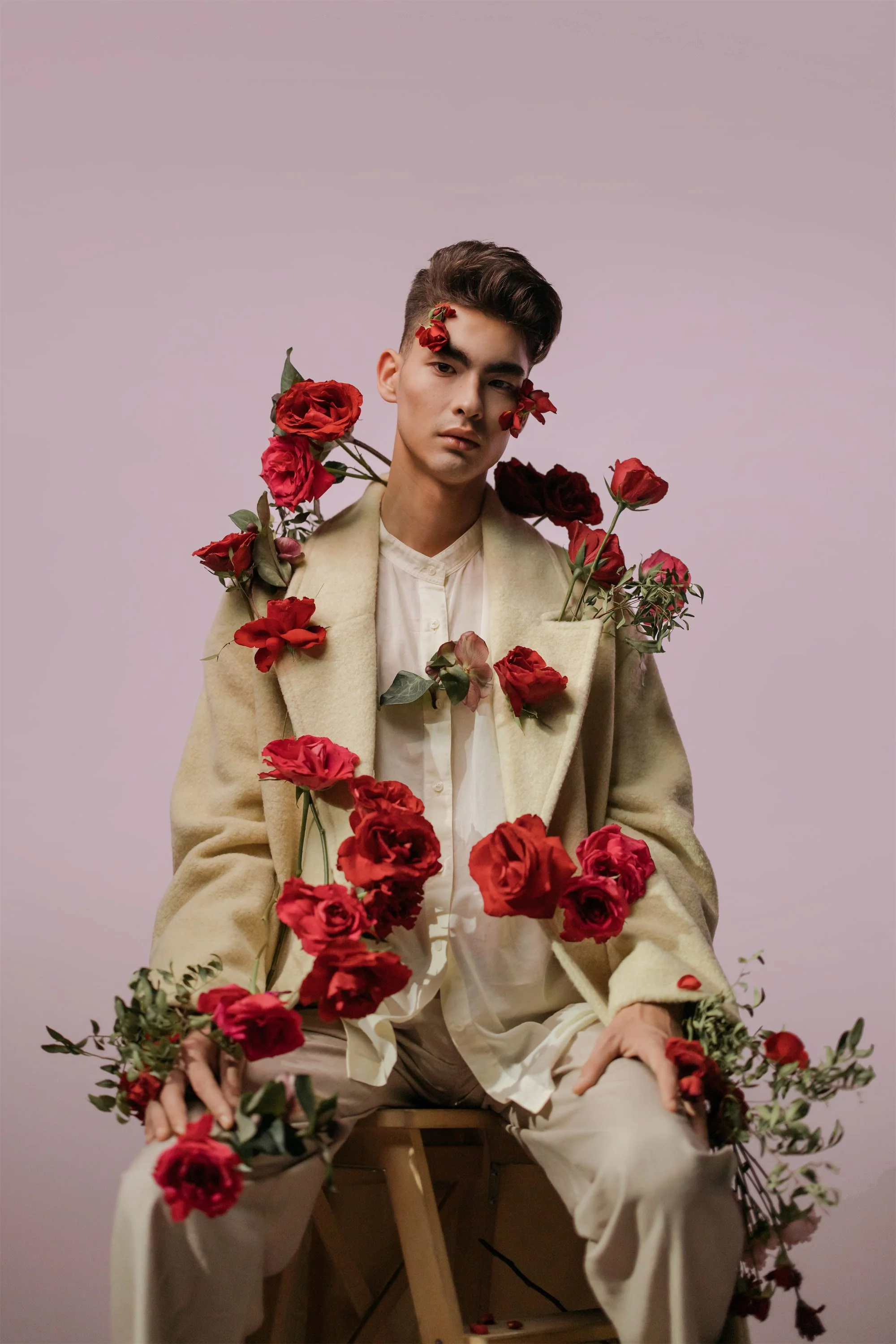 A young man sitting on a wooden stool wearing a beige coat over a white shirt, surrounded and adorned with red roses. The background is a plain light pink color.