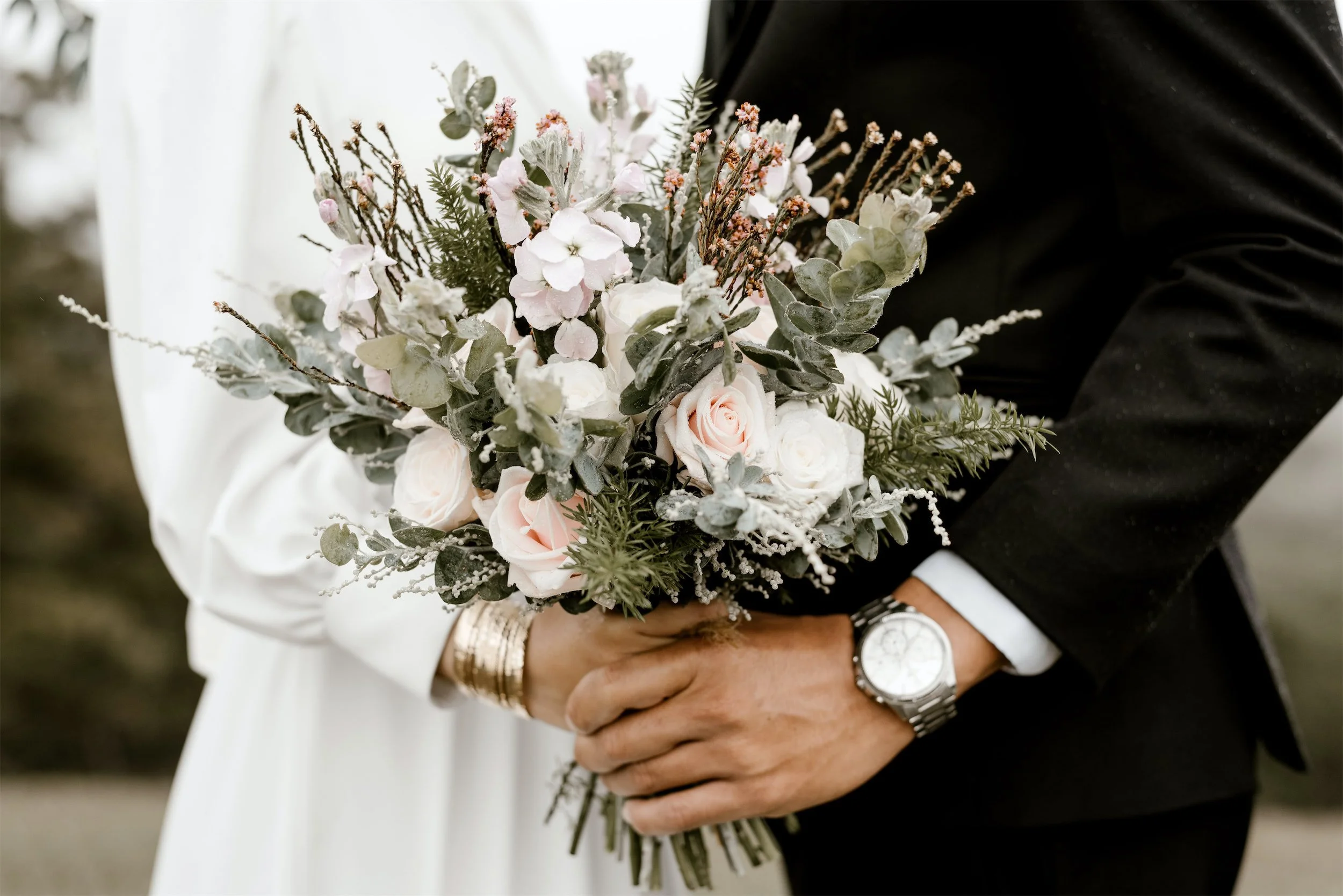 A newlywed couple holding a floral wedding bouquet. The bride's white dress is partially visible, and the groom is wearing a black suit, white shirt, and a watch. The bouquet contains pink roses, green foliage, and other delicate flowers.