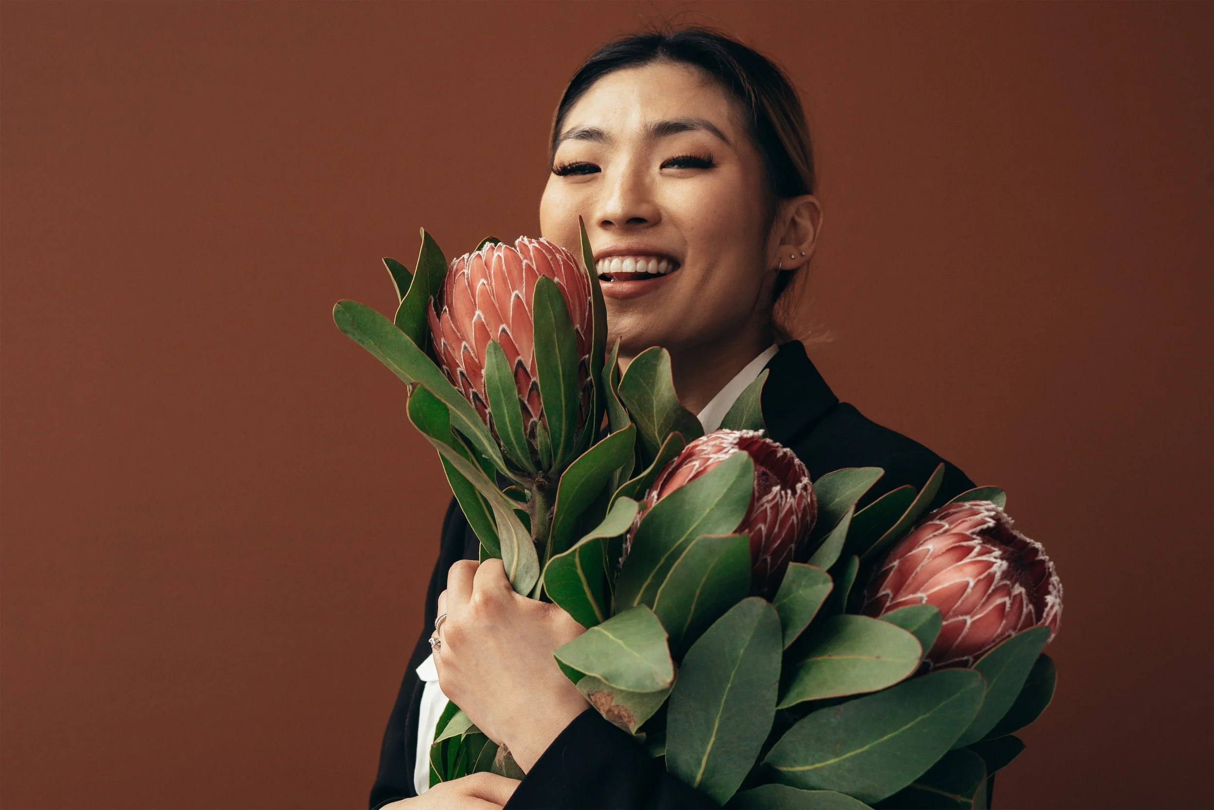 A woman smiling while holding a bouquet of large flowers with green leaves against a brown background.