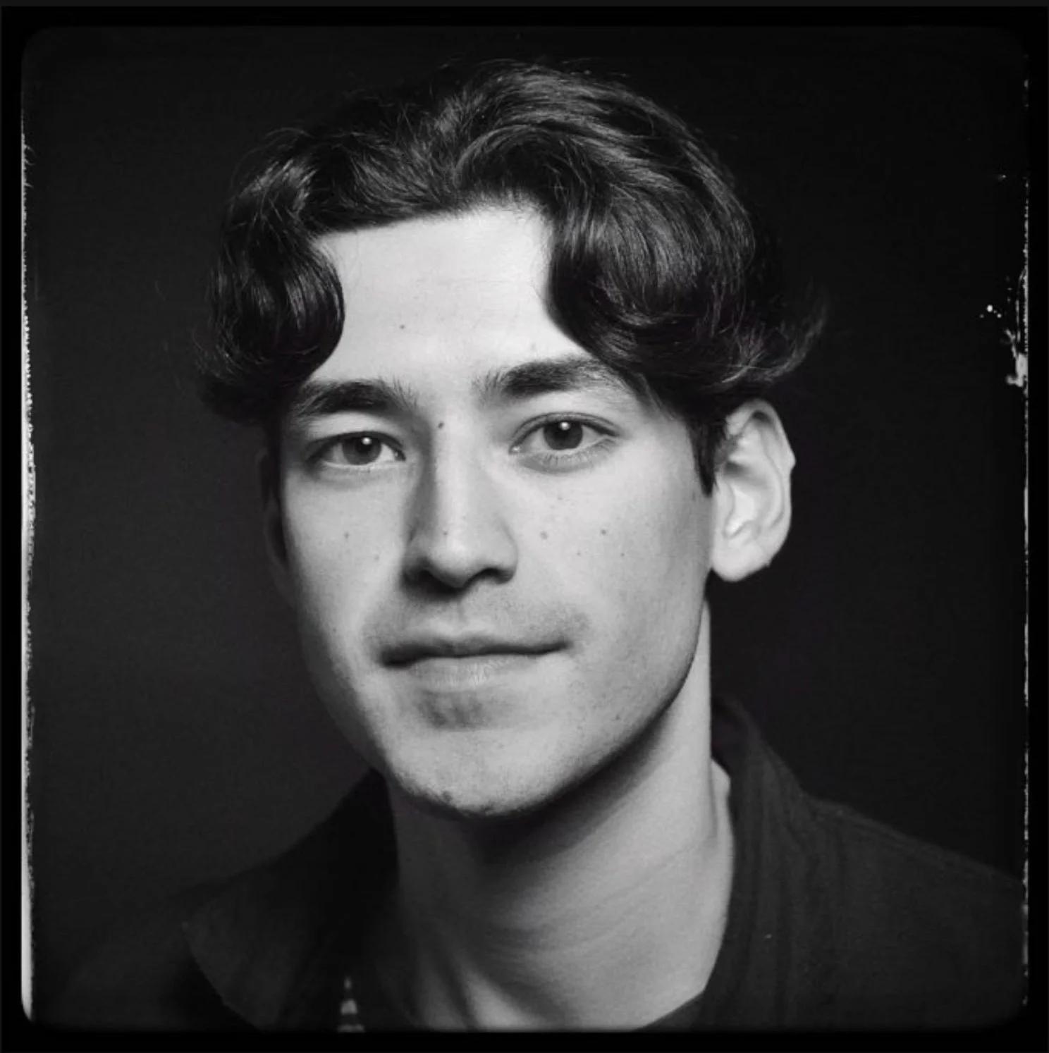 Black and white portrait of a young man with wavy dark hair, light skin, wearing a dark jacket, smiling slightly with a neutral expression, against a dark background.