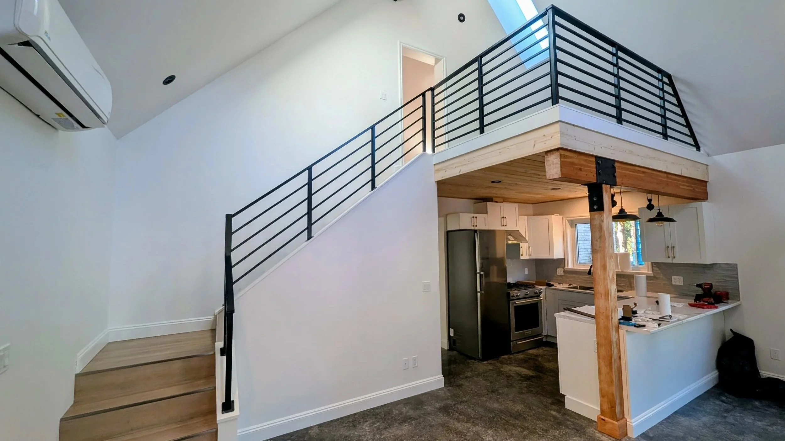 Interior view of a modern, open-concept living space with a loft. The space features a staircase with black metal railings, hardwood steps, and a wooden support beam. Visible kitchen area includes white cabinetry, stainless steel appliances, and pendant lighting. White walls and a ceiling with skylights provide natural light.