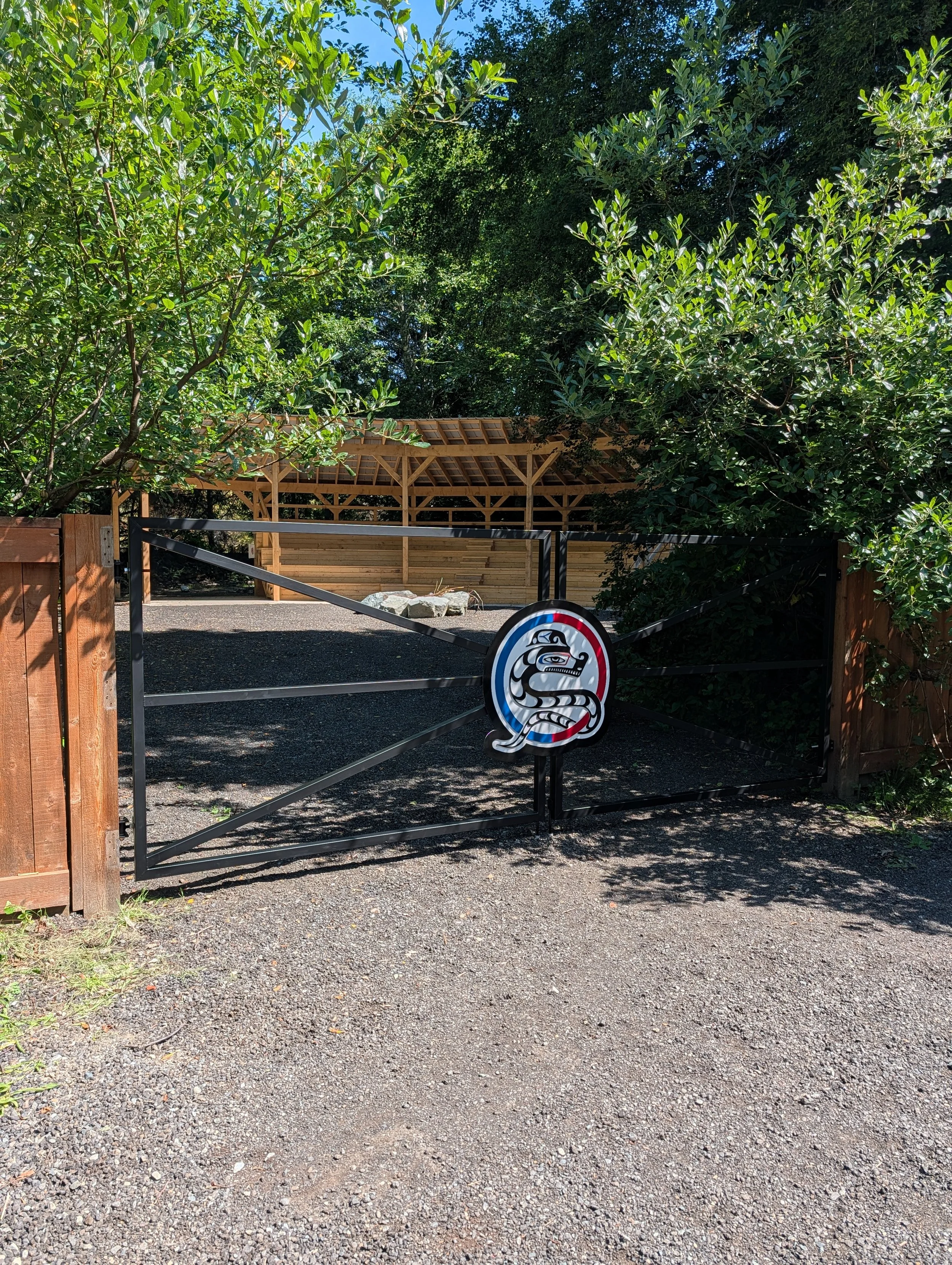 A black gate with a colorful sticker of a snake and a bird, leading to a gravel pathway surrounded by dense green trees, with a wooden structure and stone accents in the background.