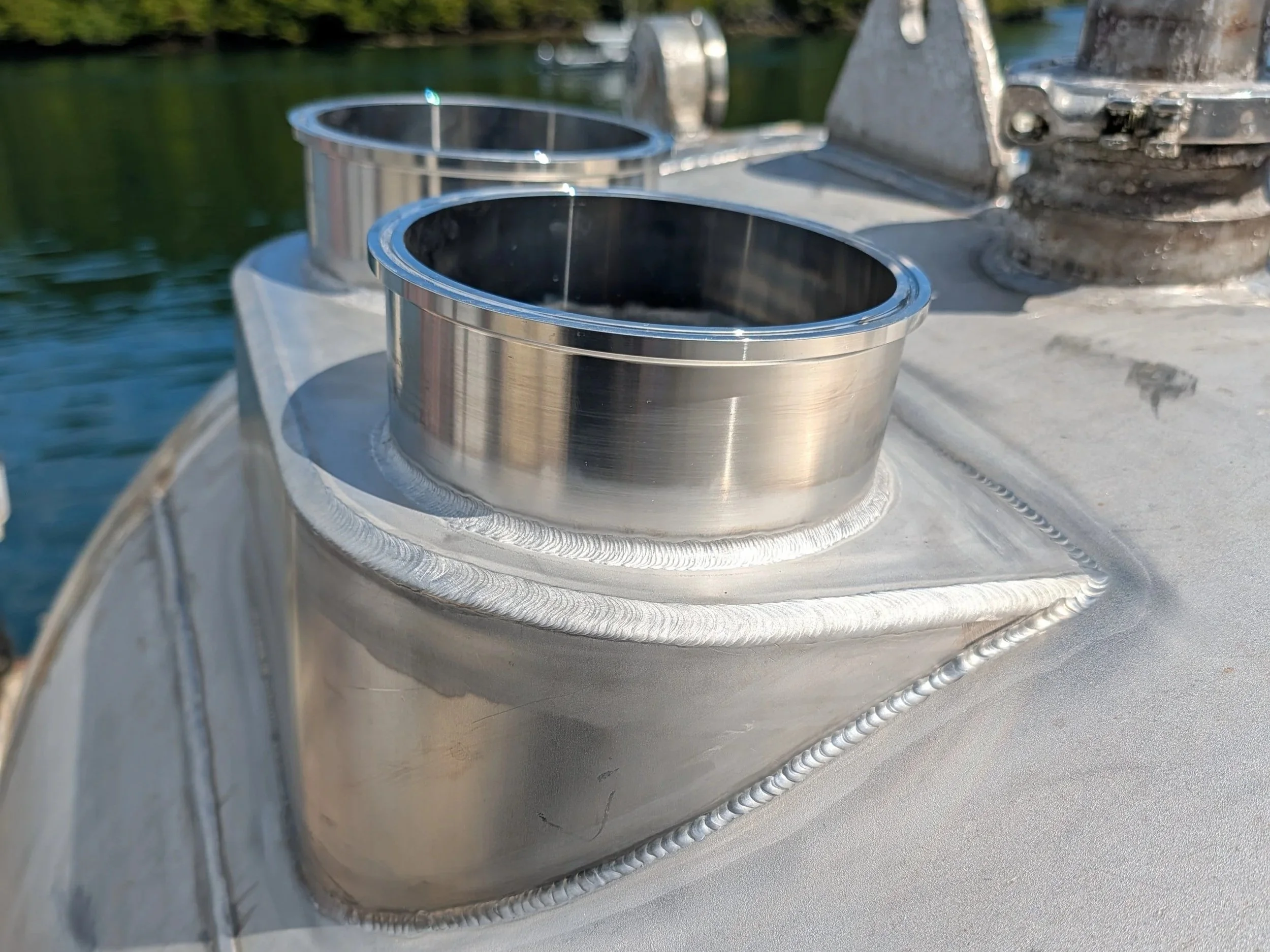 Close-up of a boat's metal hull with two circular metal fittings and a connected cylinder near the water, with trees and water in the background.