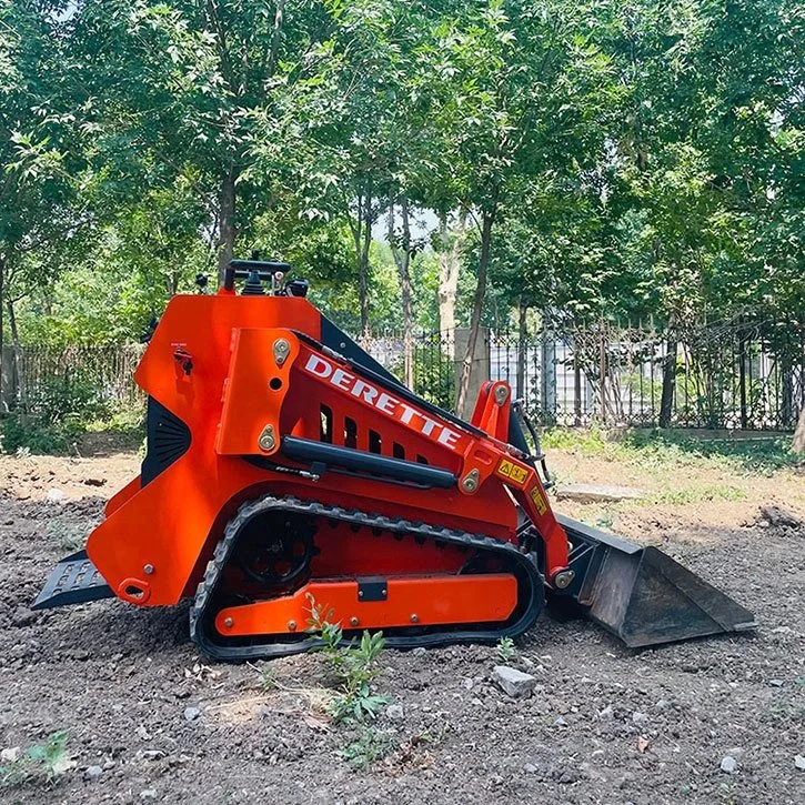 Orange mini skid steer loader parked on soil with trees in background.