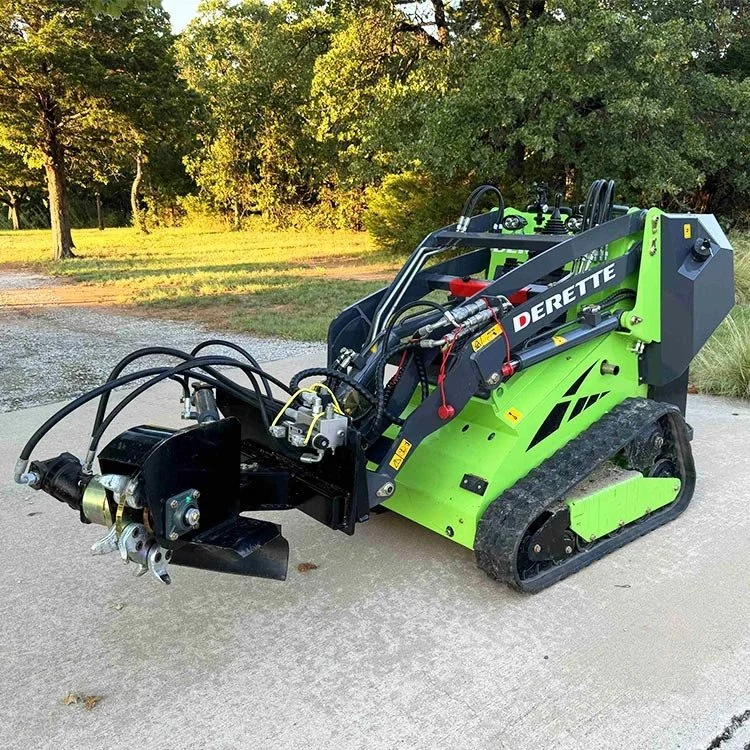 Green and black mini skid steer loader with attachments, parked on concrete with trees in the background.