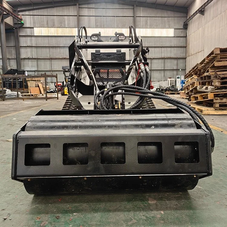 Industrial workshop with a compact tracked loader featuring an attachment, likely a mulching head, with hydraulic hoses visible.