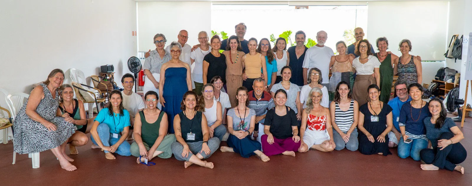 Grupo de pessoas sorrindo em uma sala, algumas sentadas no chão e outras em pé, em um ambiente bem iluminado com plantas ao fundo.