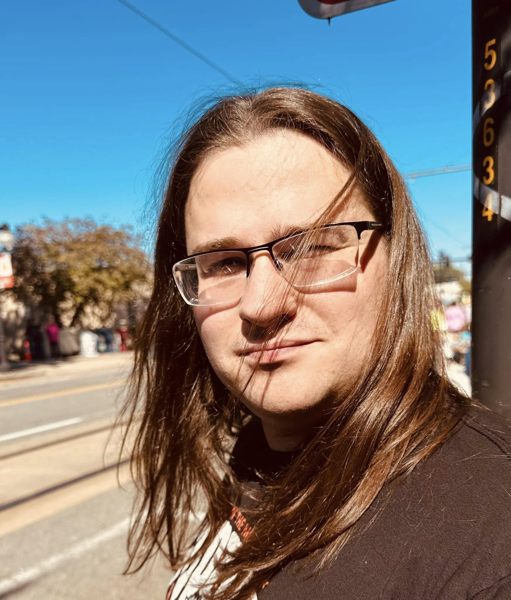 A clean shaven man with long brown hair and glasses is standing outdoors on a sunny day with a clear blue sky and trees in the background.