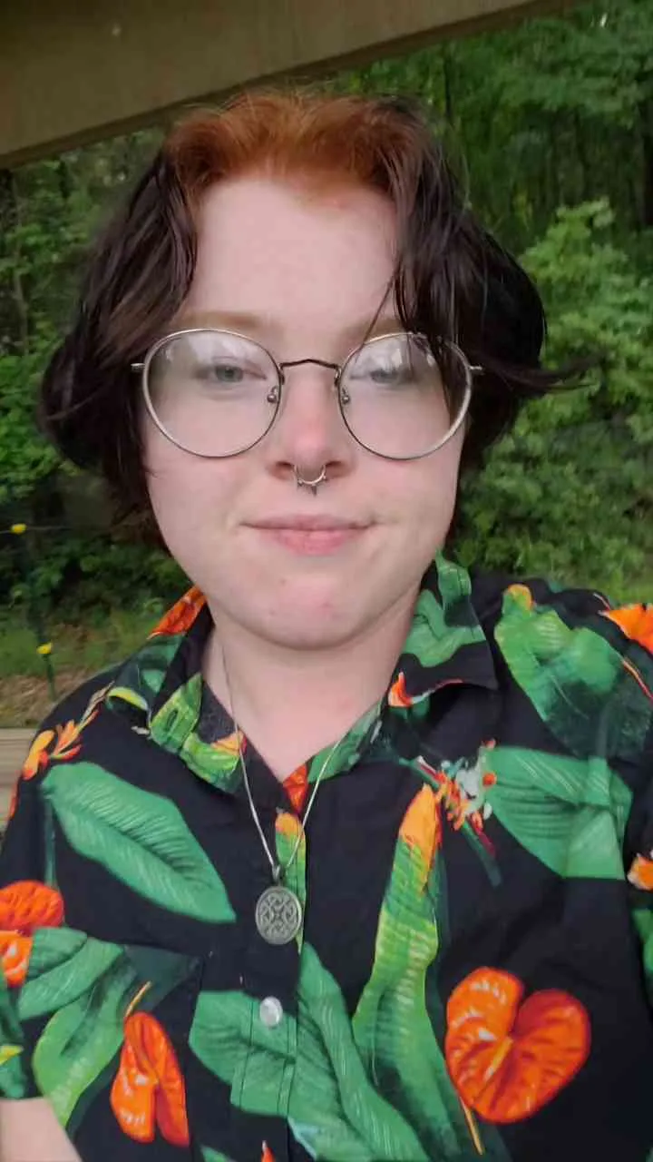 A young man person with short, two-tone hair, big round glasses, a septum piercing, and a floral Hawaiian shirt standing outdoors with green trees in the background.