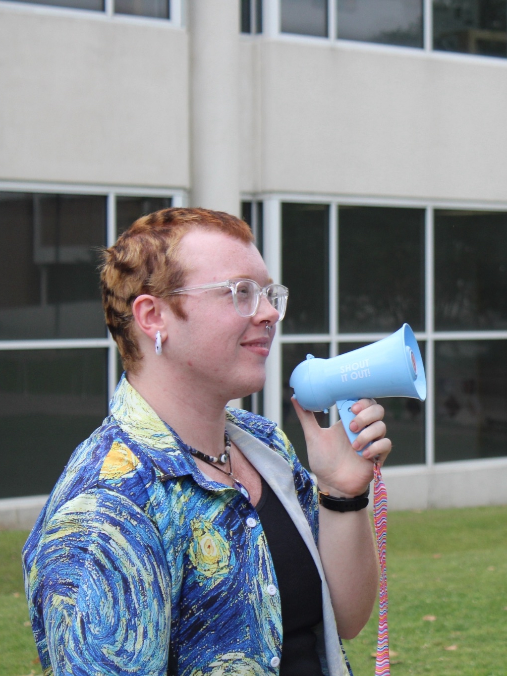A young man person with short red hair, clear framed glasses, a septum piercing and gauge earrings. He is wearing a Starry Night Van Gough shirt and carrying a small blue megaphone