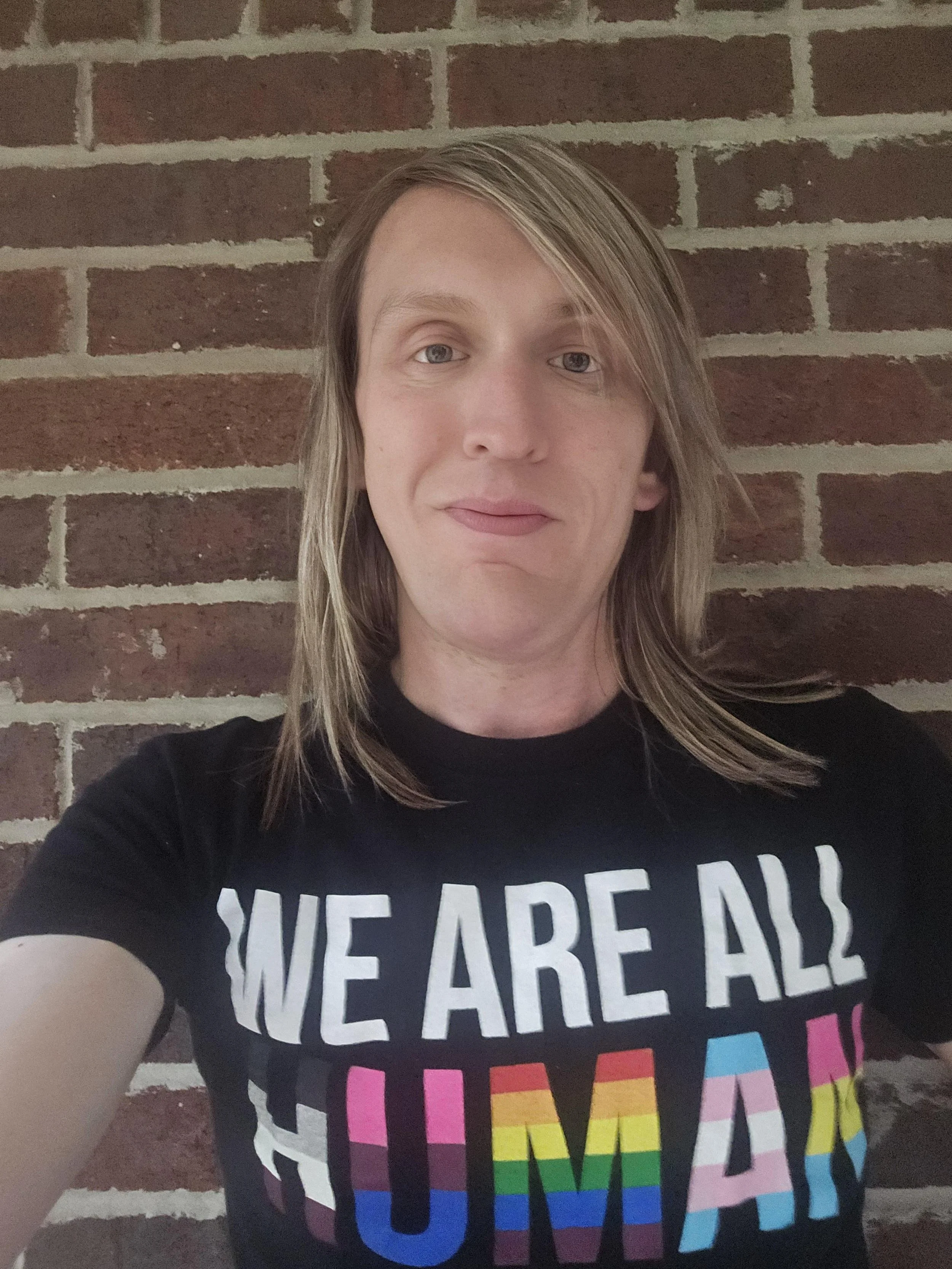 A white masculine presenting individual with long blonde hair wearing a shirt that says "We are all human"