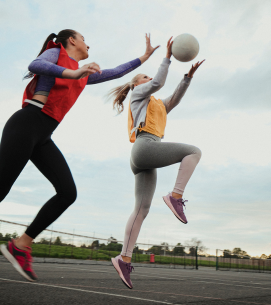 Two women playing volleyball outdoors on a cloudy day, one jumping to hit the ball while the other stands nearby.