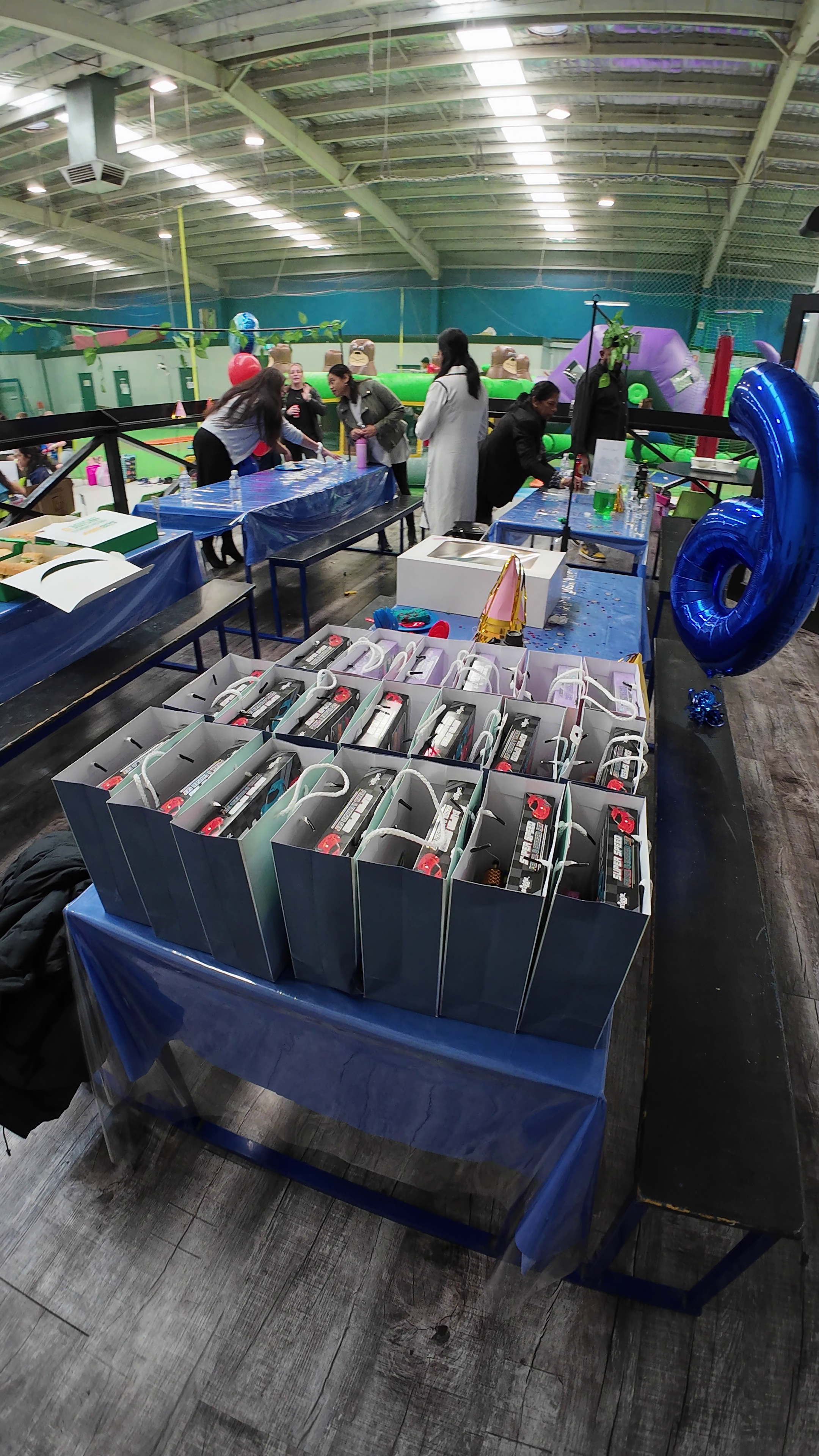 Table with blue tablecloth holding numerous electronic devices in boxes at an indoor amusement center with people setting up decorations and activities in the background.