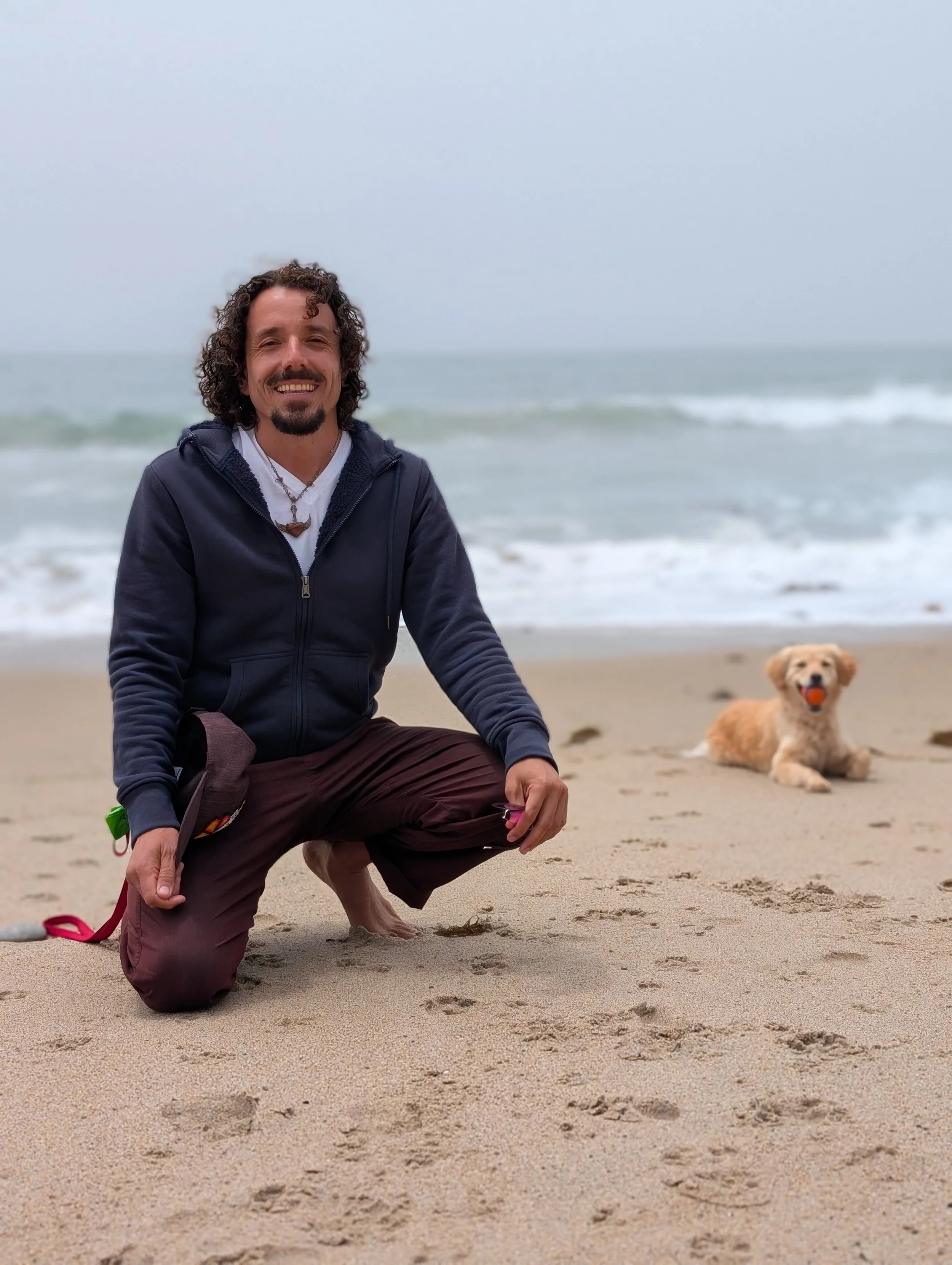A smiling man with curly hair and a beard kneeling on the sandy beach with a small dog laying nearby. The ocean waves are in the background on a cloudy day.
