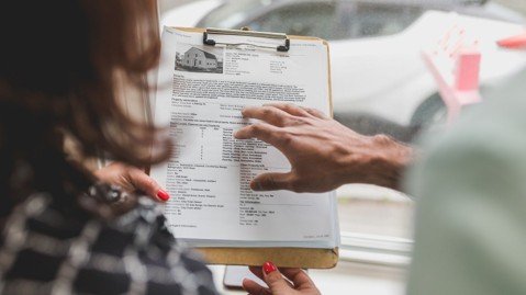 Buyer and real estate professional reviewing property documents on a clipboard near a home window.