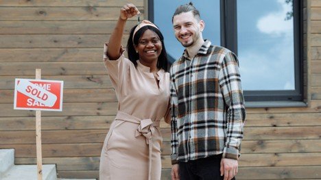 Happy homeowners holding house keys near a sold sign, representing long-term benefits of buying a home.