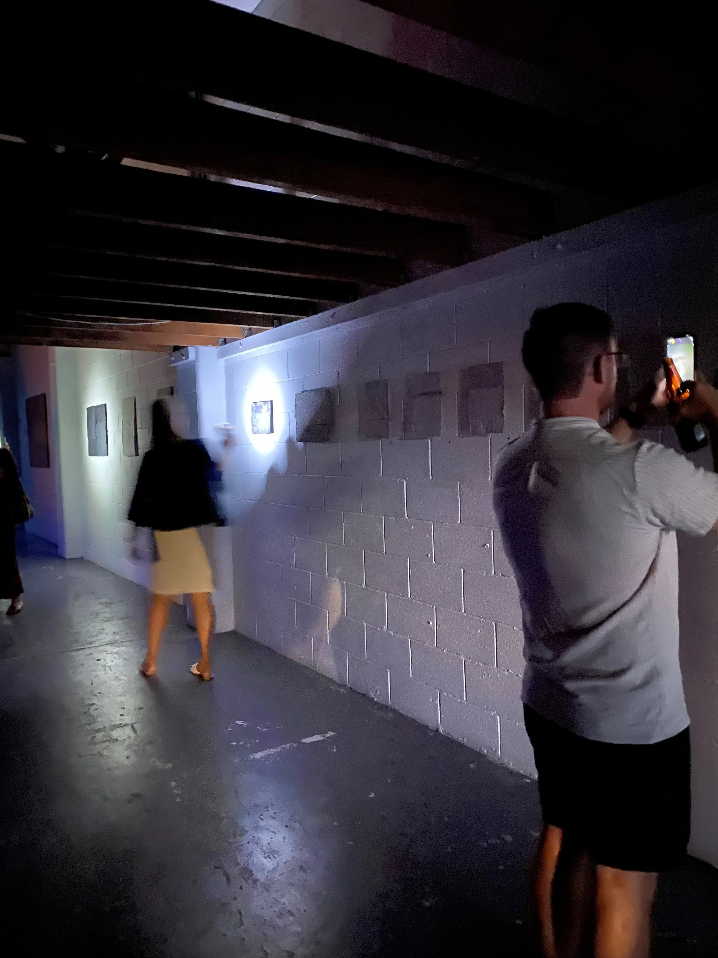 Two people looking at an exhibition at Vacant Assembly.  They are holding up their phones to see the artwork on the walls as exhibition was deliberately in the dark.