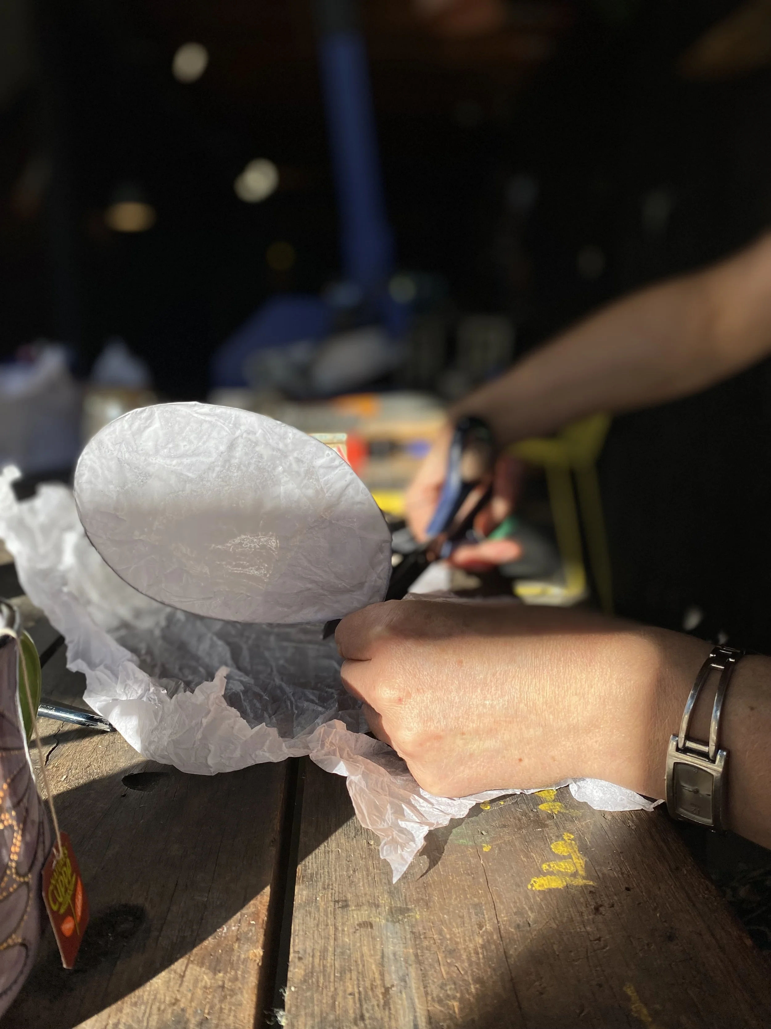 In the foreground a women is in conversation about making ceramics across a table, which is full of potters tools and some cups in their initial stages.  in the background are four women talking and laughing. 