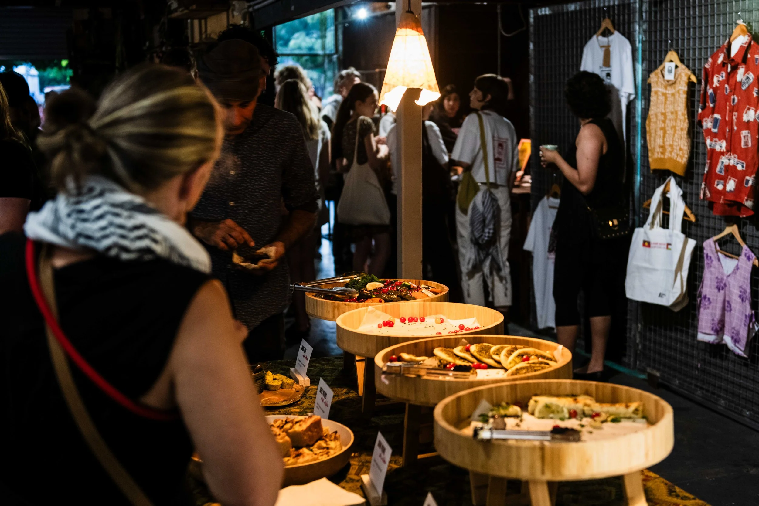 People in the foreground around big circular platters of food.  In the background are people milling around at a Vacant Assembly event.