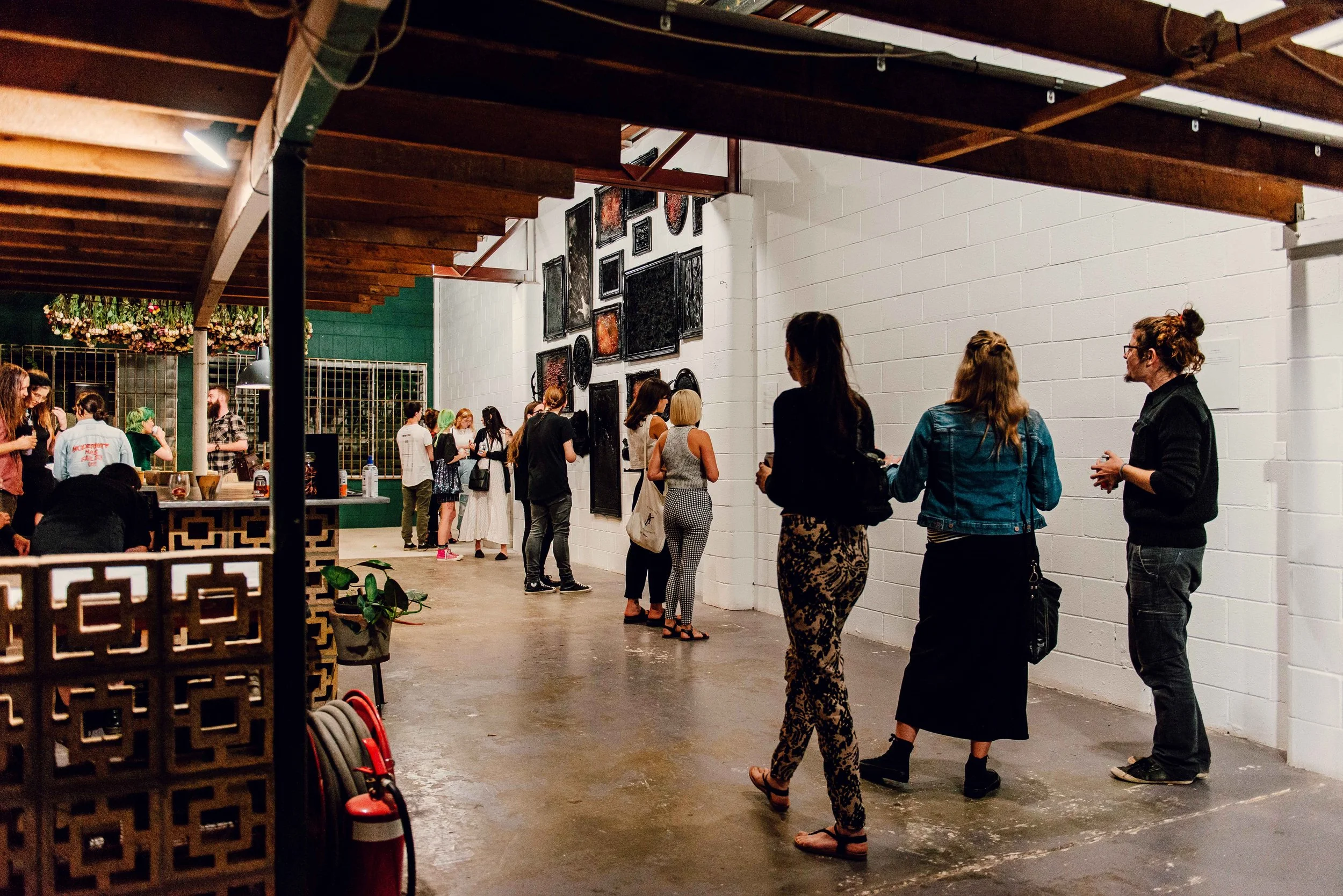 View of back wall at Vacant Assembly with a salon hanging of black or dark artworks in black frames. People are milling about at exhibition in foreground.